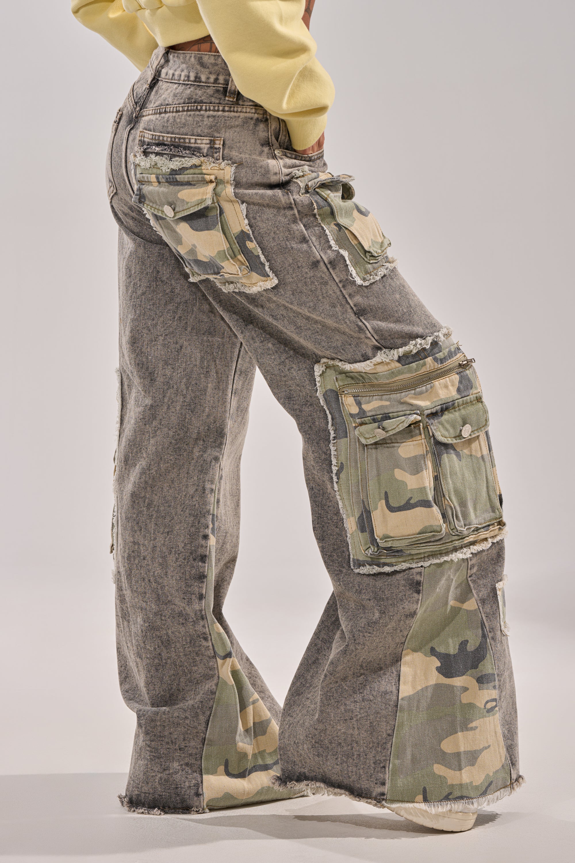 A person wears the DOWN AND DIRTY CAMO CARGO jeans—gray acid-wash denim with frayed hems, large camo cargo pockets, and unique patchwork details—styled with a light yellow cropped top and photographed from the side. 3
