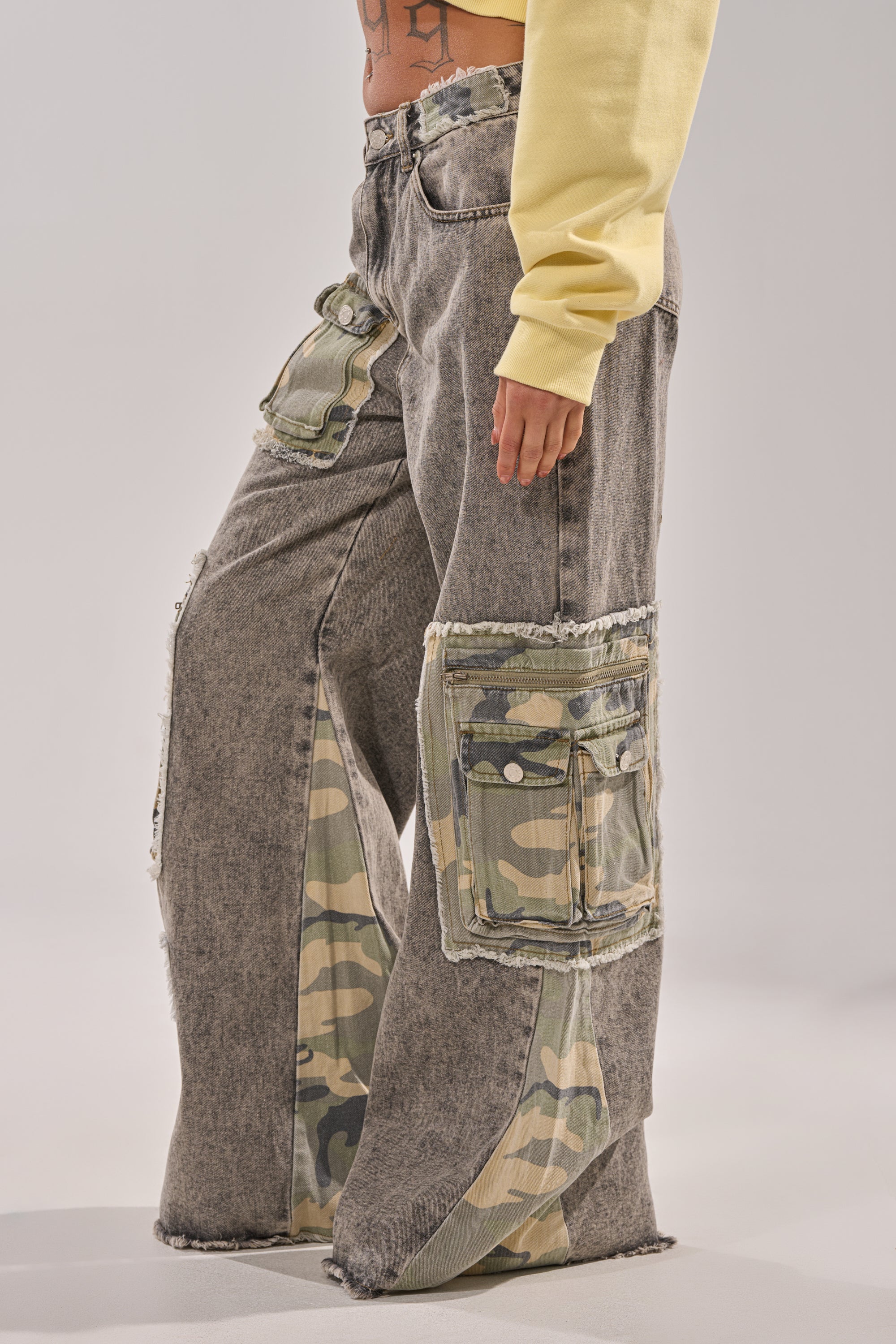 A side-view photo shows a person wearing the DOWN AND DIRTY CAMO CARGO: wide-leg, acid-wash gray denim jeans with large camo cargo pockets and frayed edges, paired with a yellow long-sleeve top. Lower torso and legs are visible. 4