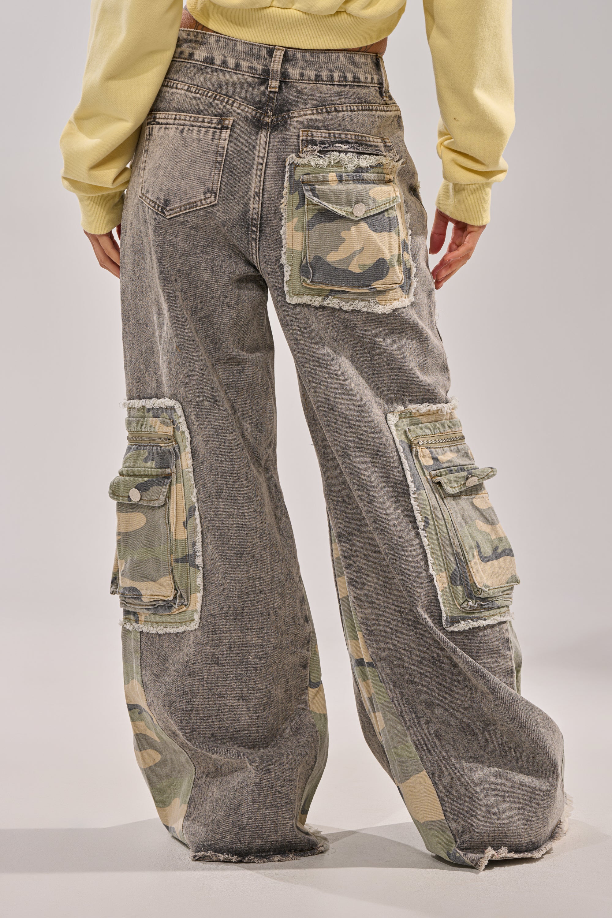 A person wearing the DOWN AND DIRTY CAMO CARGO jeans, featuring a loose flared fit with camo cargo pockets and back patches, is paired with a yellow sweatshirt. Photo taken from behind highlights the pocket details. 5