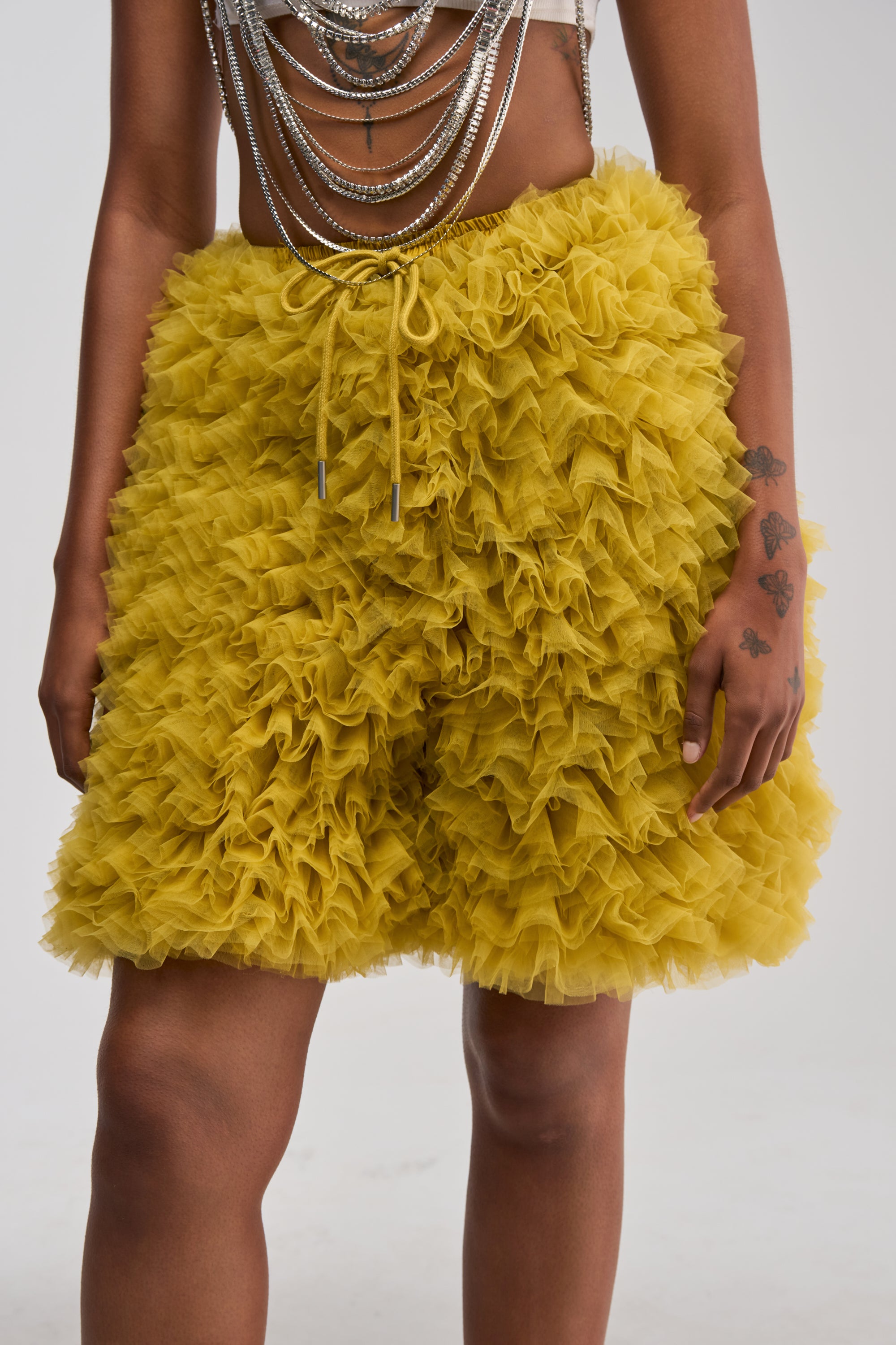 A person wearing a white top paired with the SOMETHING CRAZY RUFFLE SHORT in chartreuse and layered silver chains, with tattoos visible on one arm, standing against a plain background.