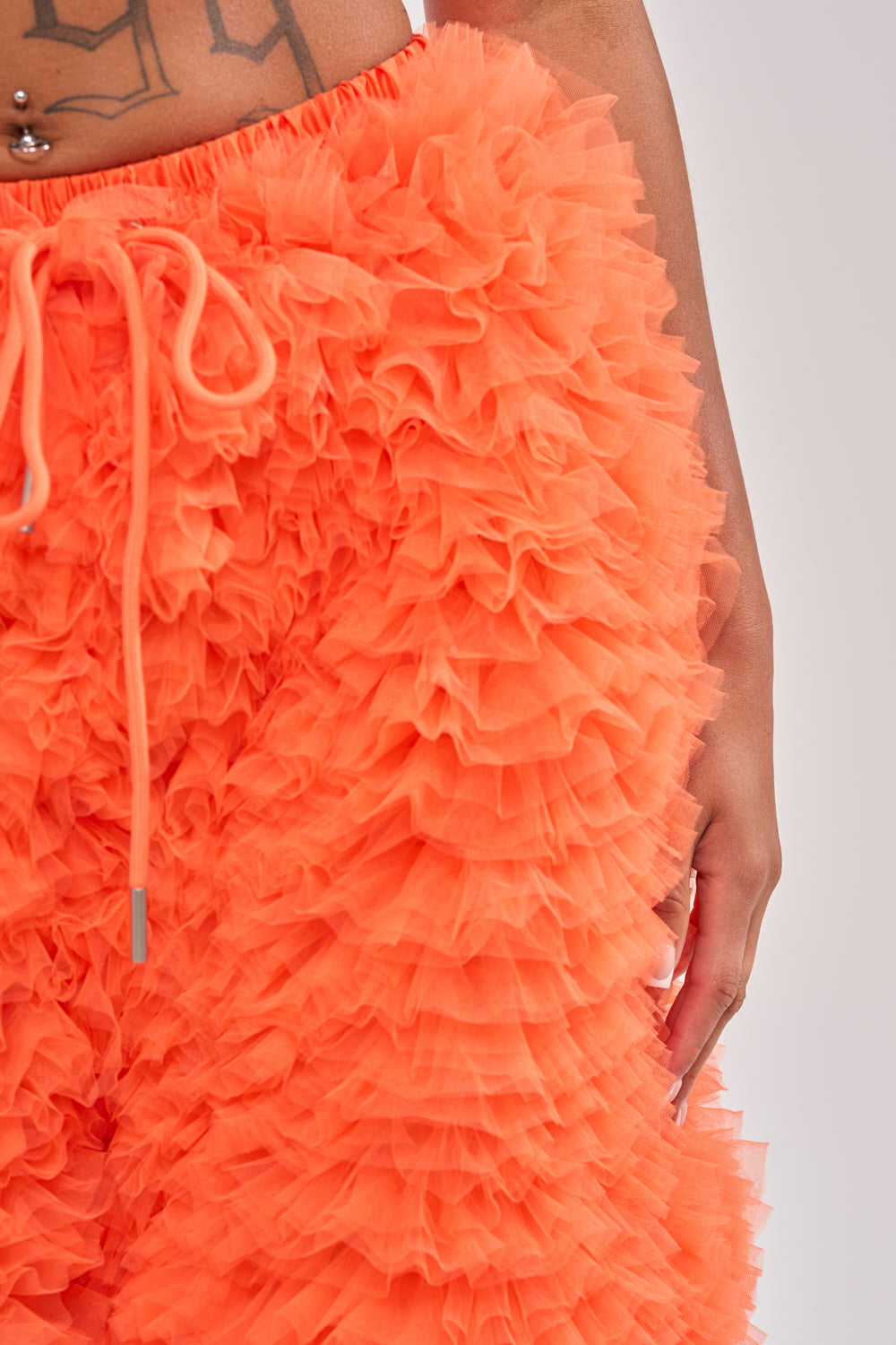 Wearing the SOMETHING CRAZY RUFFLE SHORT in bright orange with a drawstring waist, the image focuses on their midsection, displaying tattoos, a belly button piercing, and one hand at their side. 6