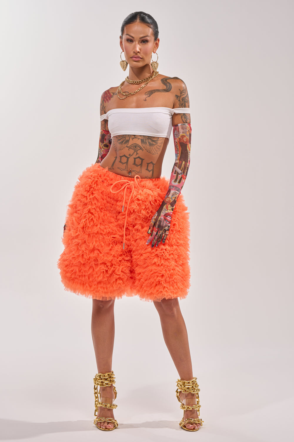 A tattooed woman poses confidently in a white bandeau top, gold accessories, gold heels, and the SOMETHING CRAZY RUFFLE SHORT in vibrant orange against a plain background. 7