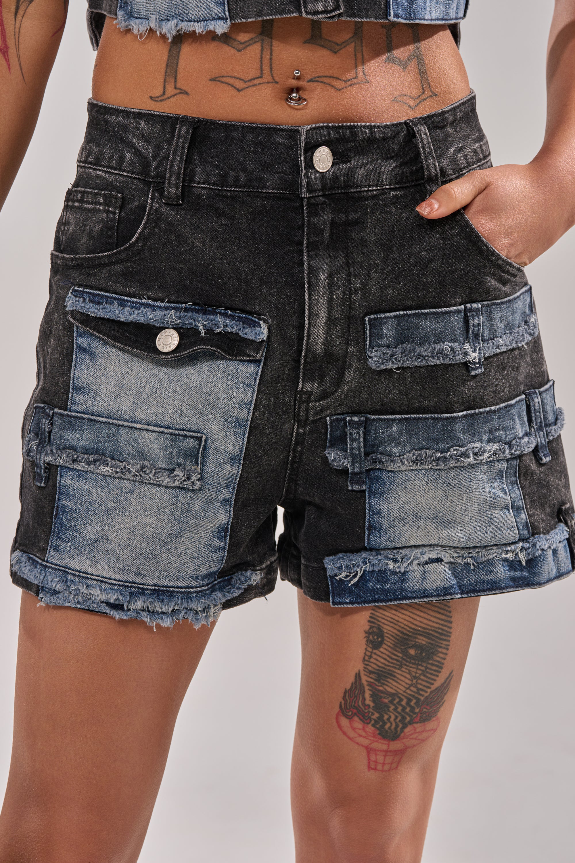 A person wears the RAJ DENIM SHORT, featuring a high-rise waist, frayed hems, blue denim patch pockets, and decorative strips, paired with a matching crop top and showcasing tattoos on their thigh, stomach, and arms. 0