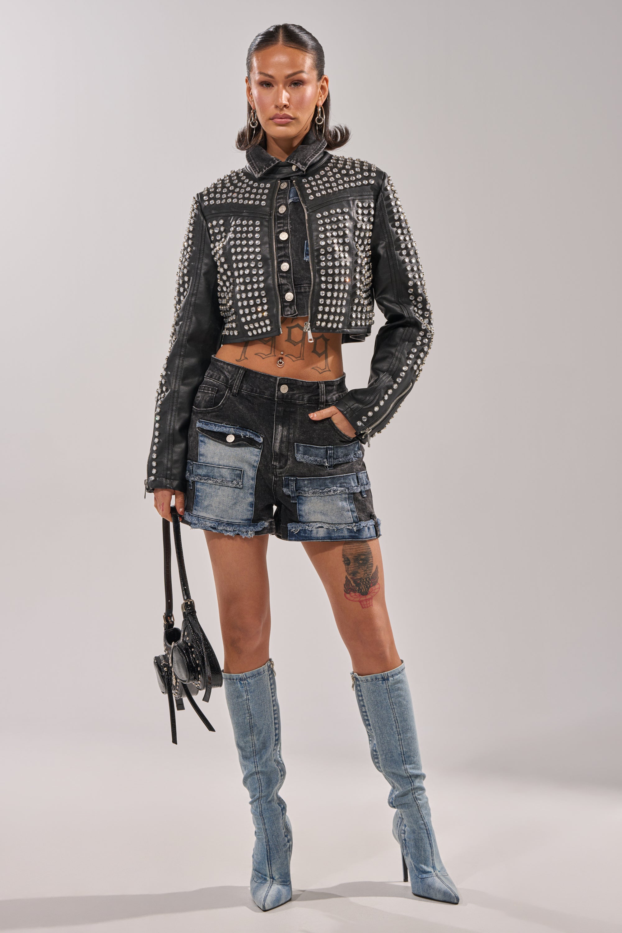 A woman with tattoos and pigtails stands confidently in a studded black cropped jacket, RAJ DENIM SHORT high-rise patchwork shorts, light blue knee-high boots, and a small handbag. 3