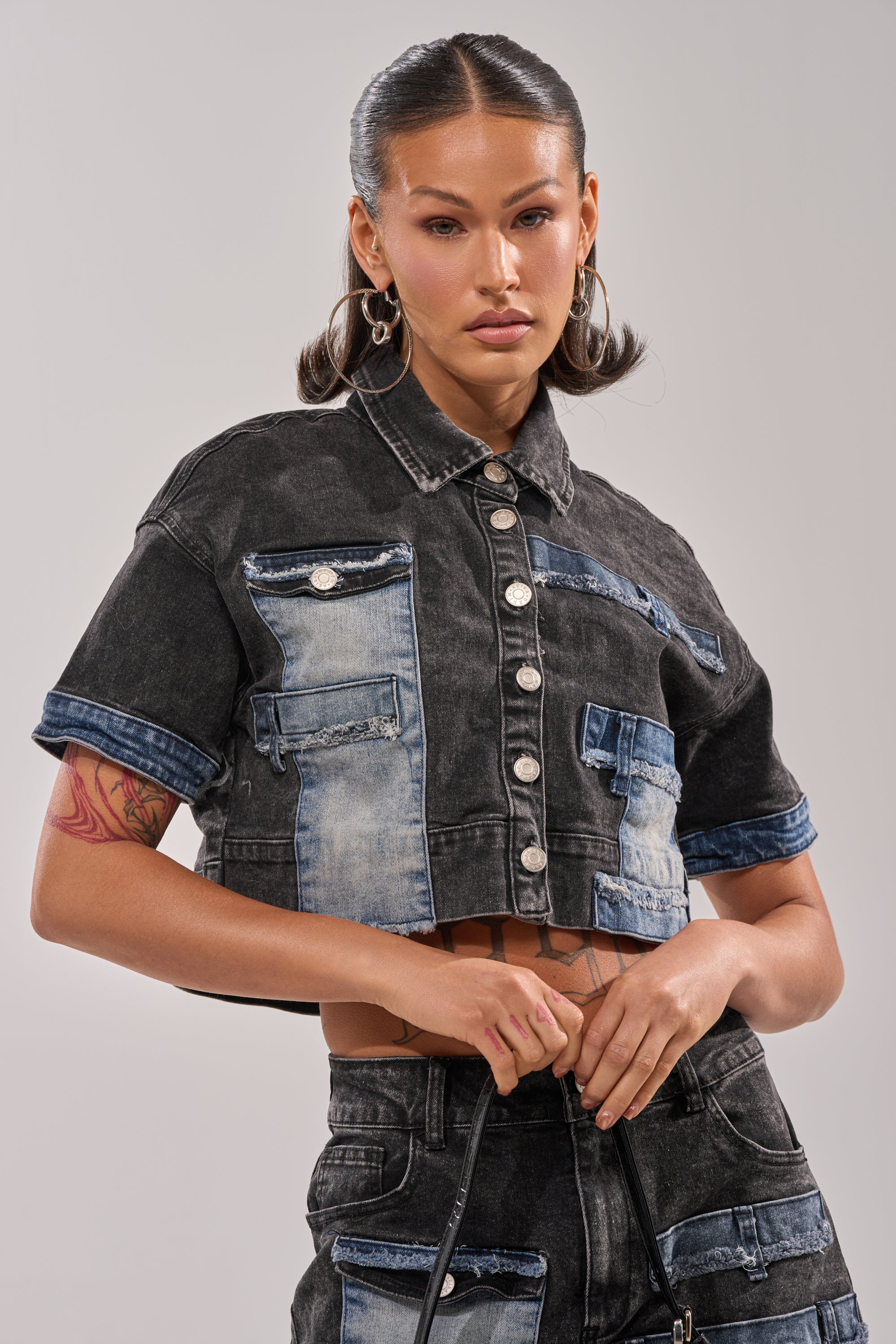 A woman with slicked-back hair wears the RAJ DENIM BUTTON DOWN and high-waisted patchwork jeans, creating a boxy silhouette. She stands confidently, holding a strap, against a light neutral background. 0