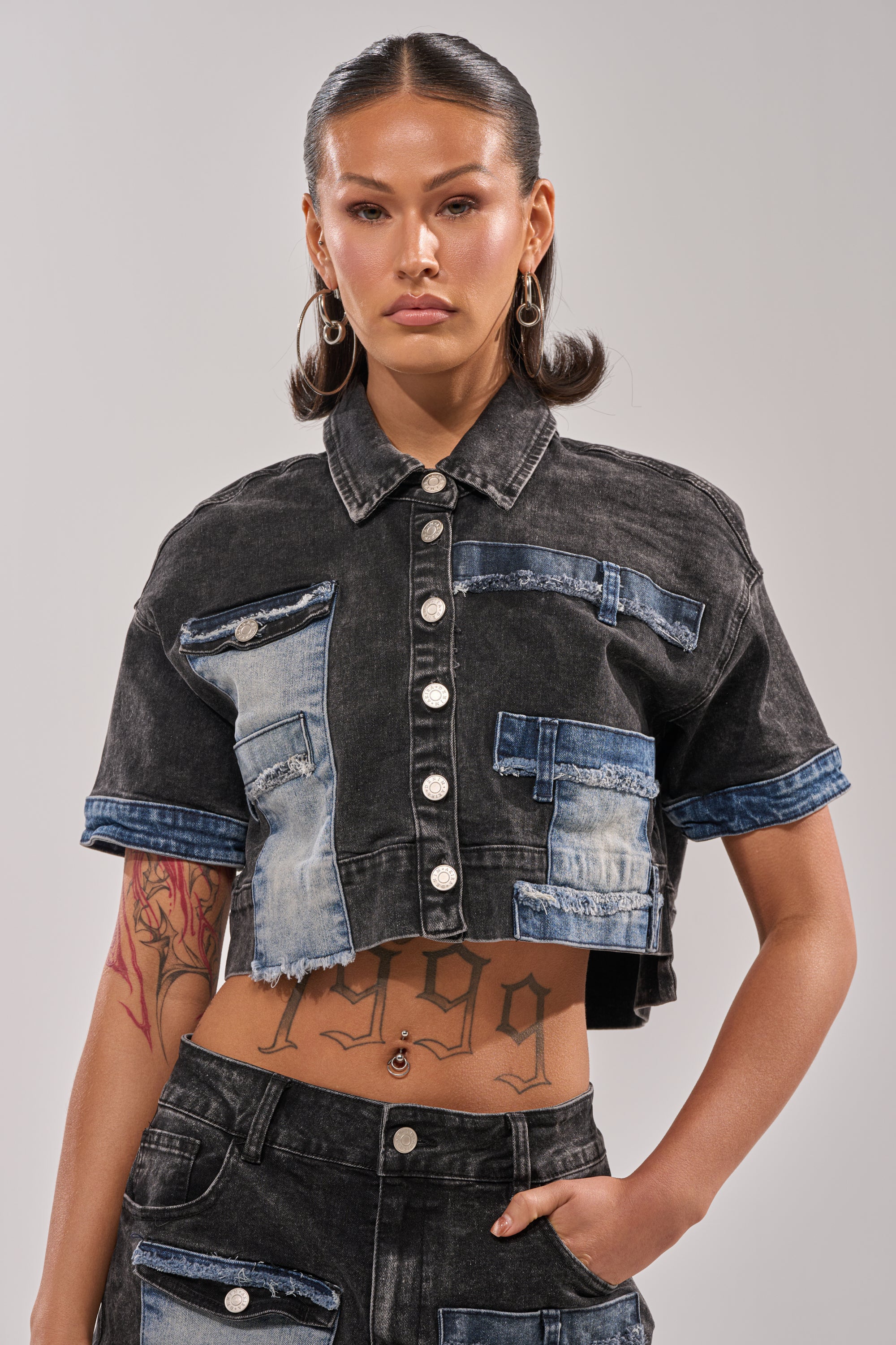 A person with slicked-back hair rocks streetwear style in the RAJ DENIM BUTTON DOWN, a cropped top with distressed patchwork, paired with matching high-waisted jeans. Their tattooed arms, midriff, and large hoop earrings complete the look. 1