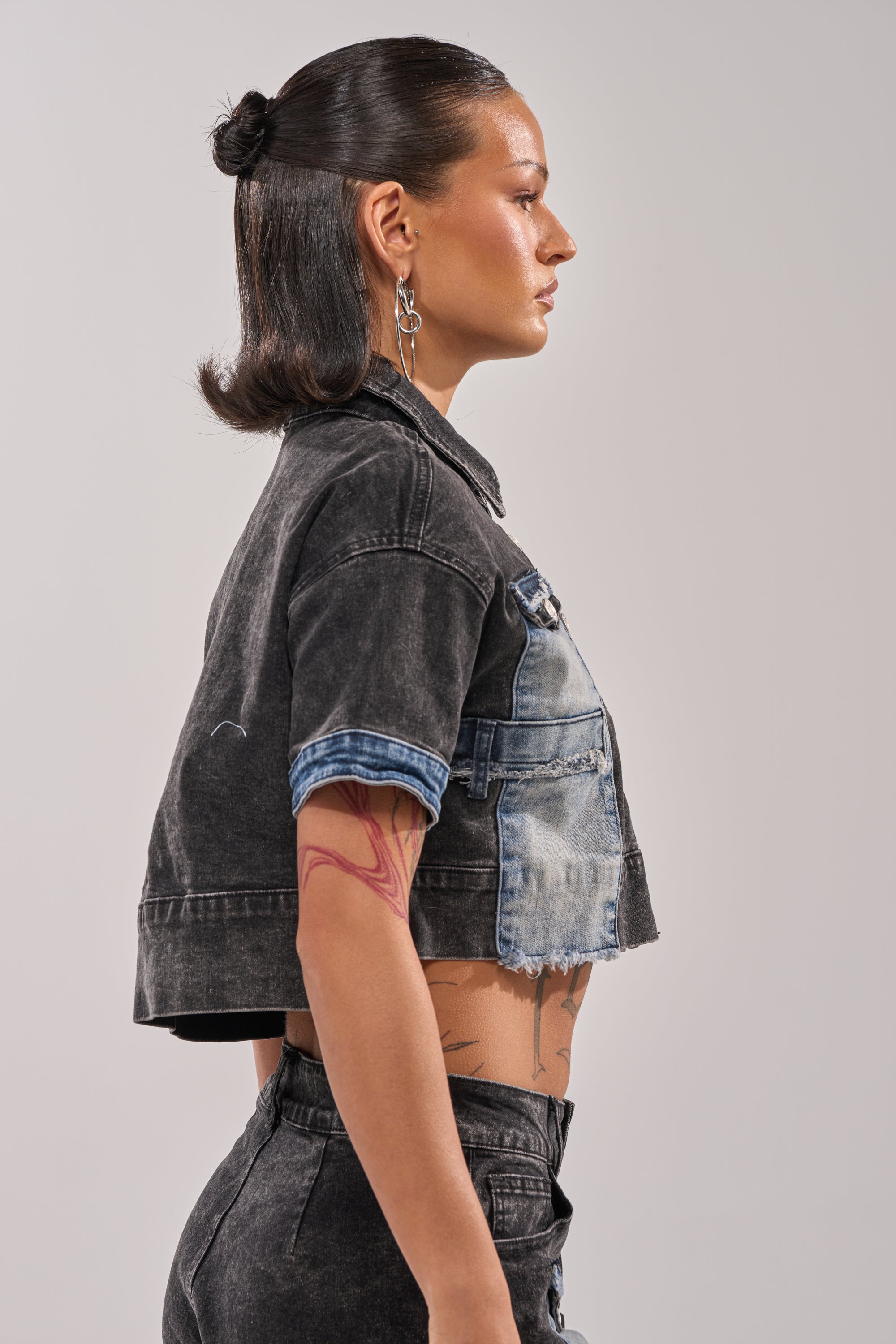 A person with straight dark hair in a clip wears the RAJ DENIM BUTTON DOWN and matching distressed patchwork denim pants, standing in profile against a plain background and displaying tattoos on their arm and torso. 2