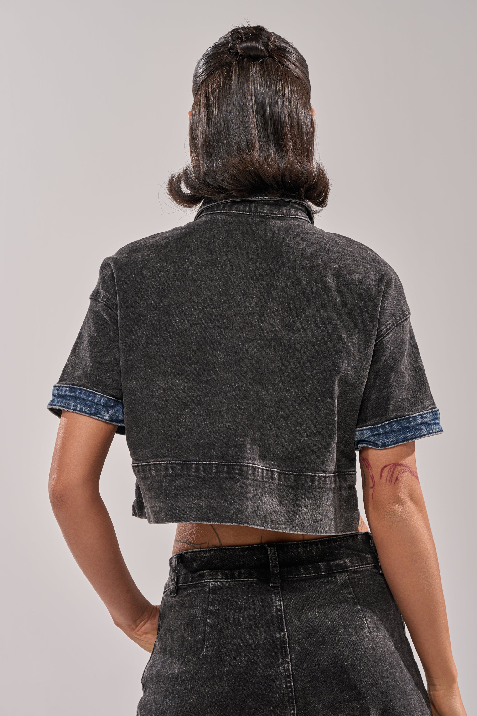 A person with shoulder-length dark hair, seen from behind, wears the RAJ DENIM BUTTON DOWN with rolled-up sleeves and matching high-waisted distressed patchwork denim pants. A tattoo on their upper arm adds an edge to their streetwear style. 4