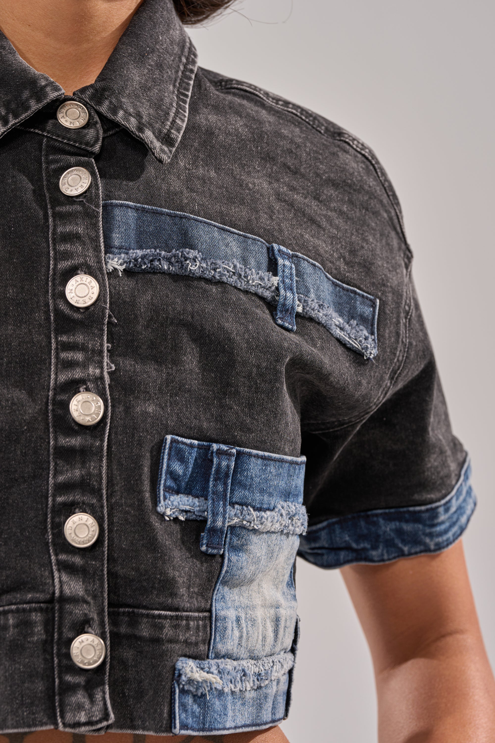A close-up of a person wearing the RAJ DENIM BUTTON DOWN, featuring frayed blue distressed patchwork accents, silver buttons, cropped fit, and rolled sleeves. Only the torso and part of one arm are visible against a light background. 5