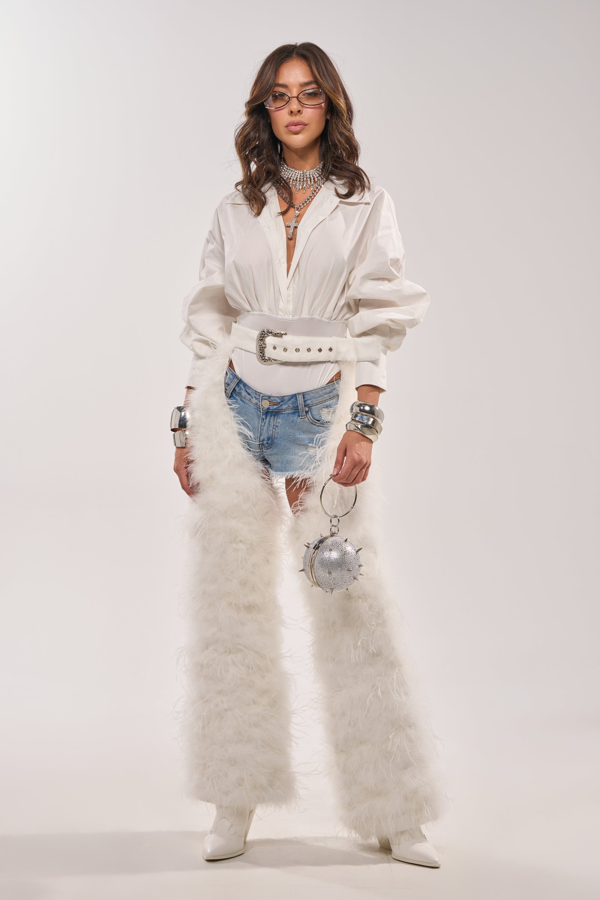 A woman poses in IM PERFECT FEATHER CHAPS, styled with a white blouse, large white belt, ripped denim shorts, white boots, layered silver jewelry, bracelets, glasses, and a silver spherical purse. 7