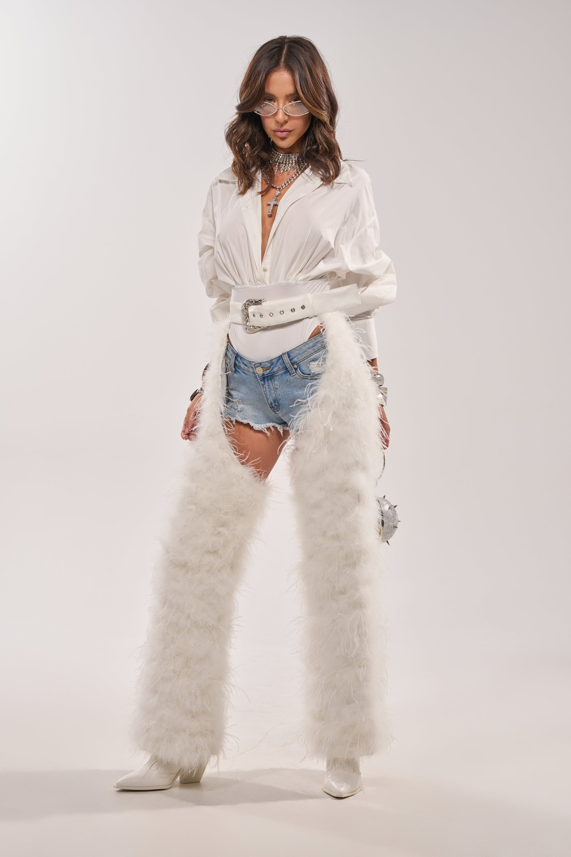 A woman poses confidently against a plain background wearing IM PERFECT FEATHER CHAPS over denim shorts, a white shirt, white boots, a white belt bag, layered necklaces, and sunglasses. 9