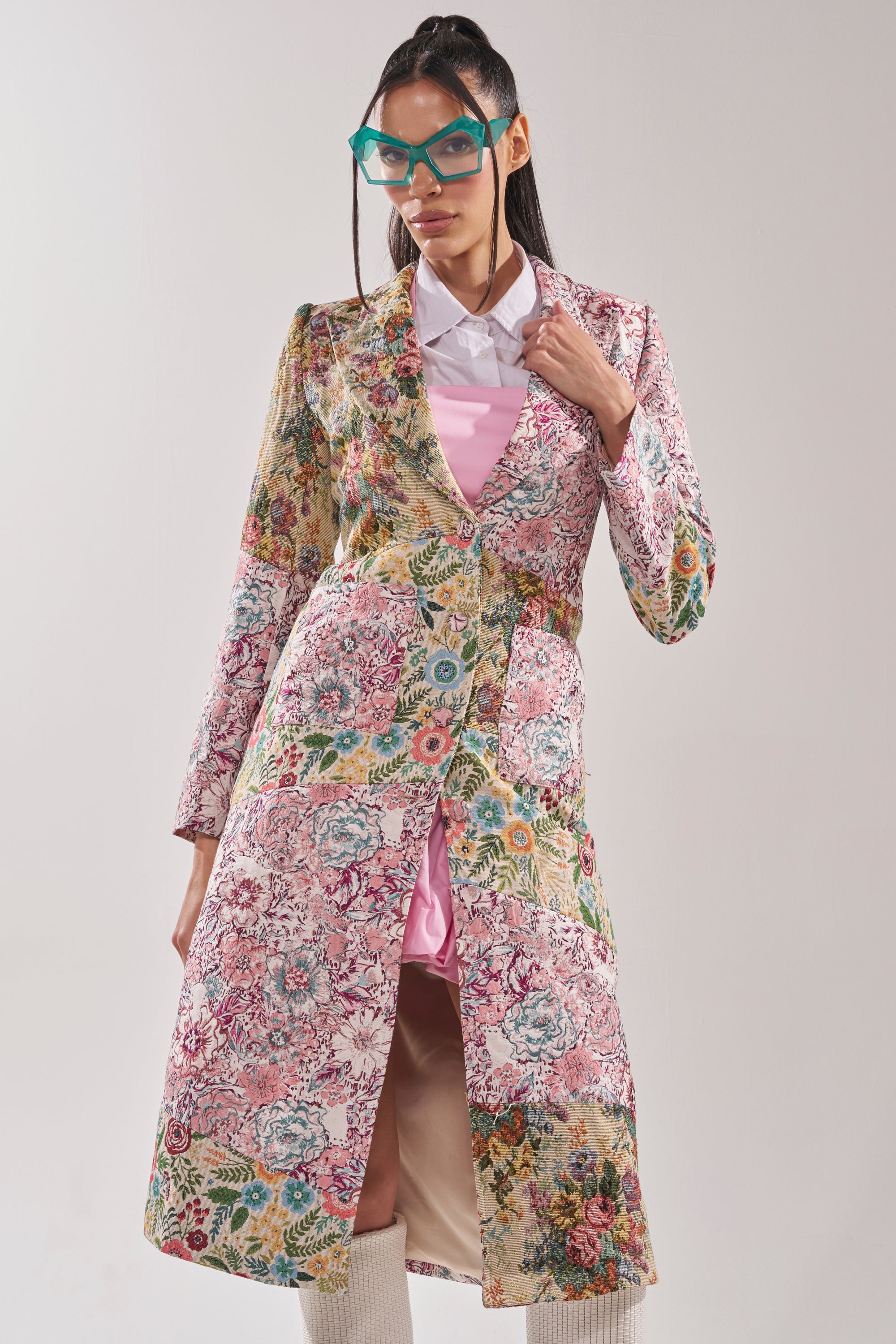 A woman models the HAD ME AT HELLO PATCHWORK TRENCH over a pink dress, with white knee-high boots and teal cat-eye glasses, standing against a plain light background. 0