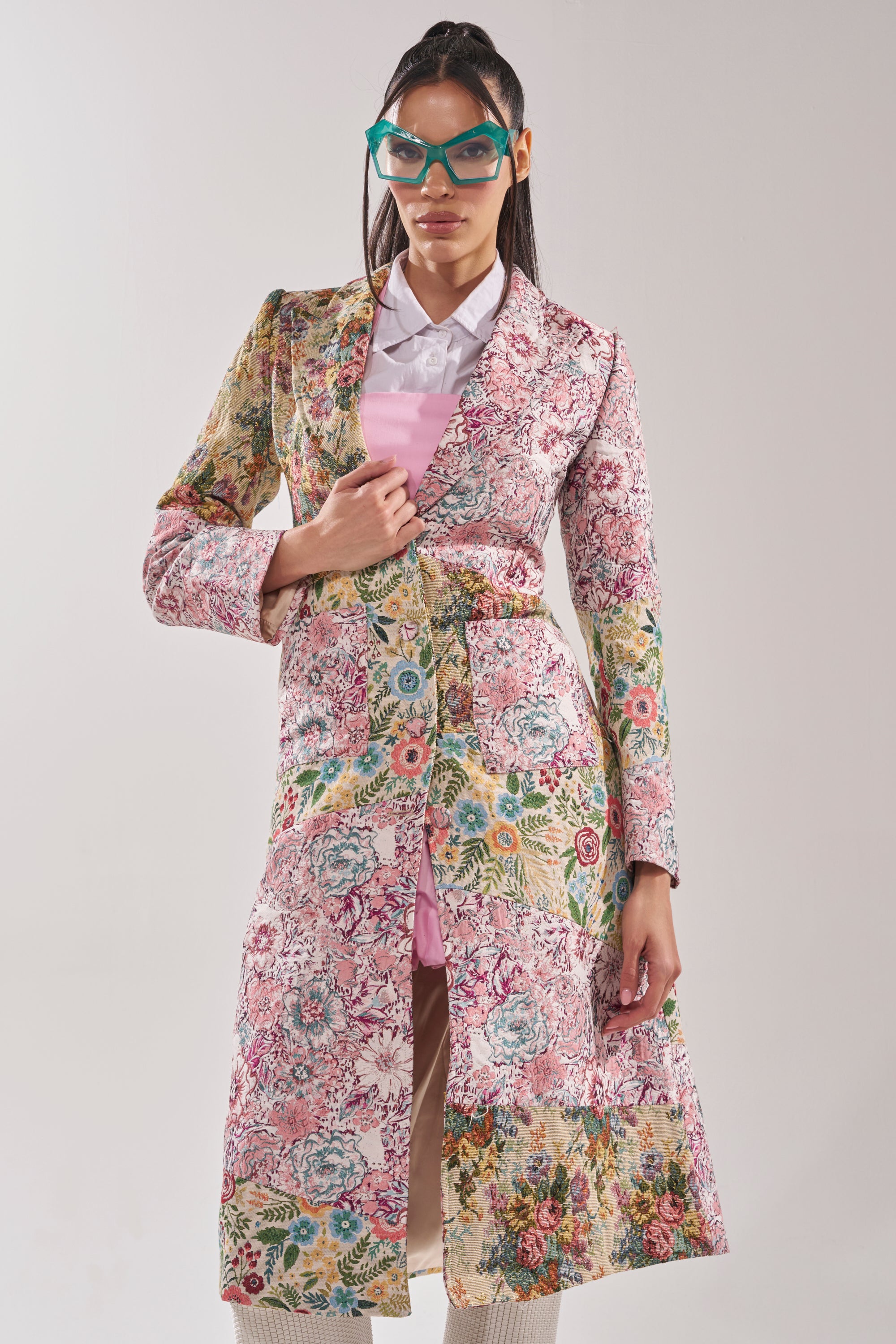 A woman wears the HAD ME AT HELLO PATCHWORK TRENCH over a light pink dress and white collared shirt, accessorized with green cat-eye glasses. The background is plain and light colored. 9