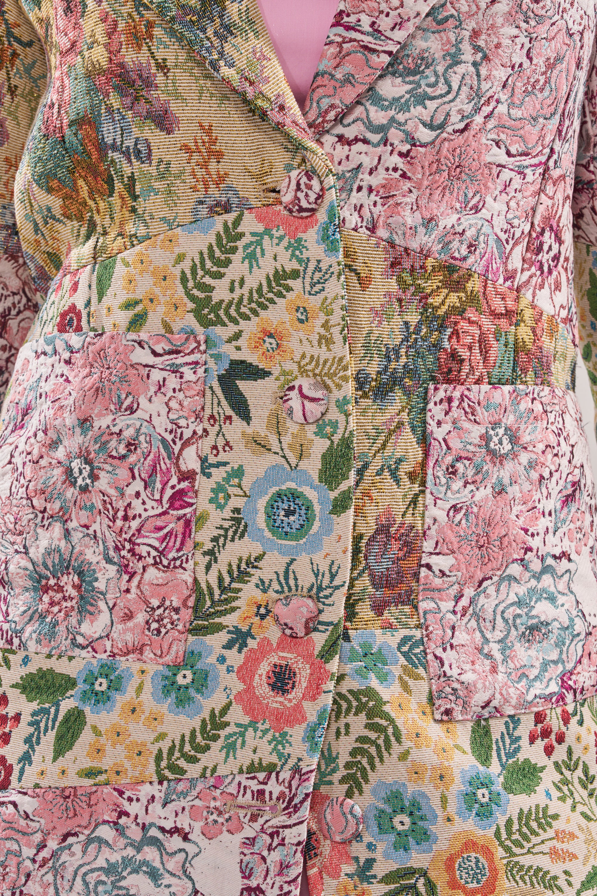 A close-up of the HAD ME AT HELLO PATCHWORK TRENCH: a colorful, vintage-inspired floral jacquard coat with a pink, yellow, green, and blue patchwork pattern, two large front pockets, and styled with a light pink top. 10