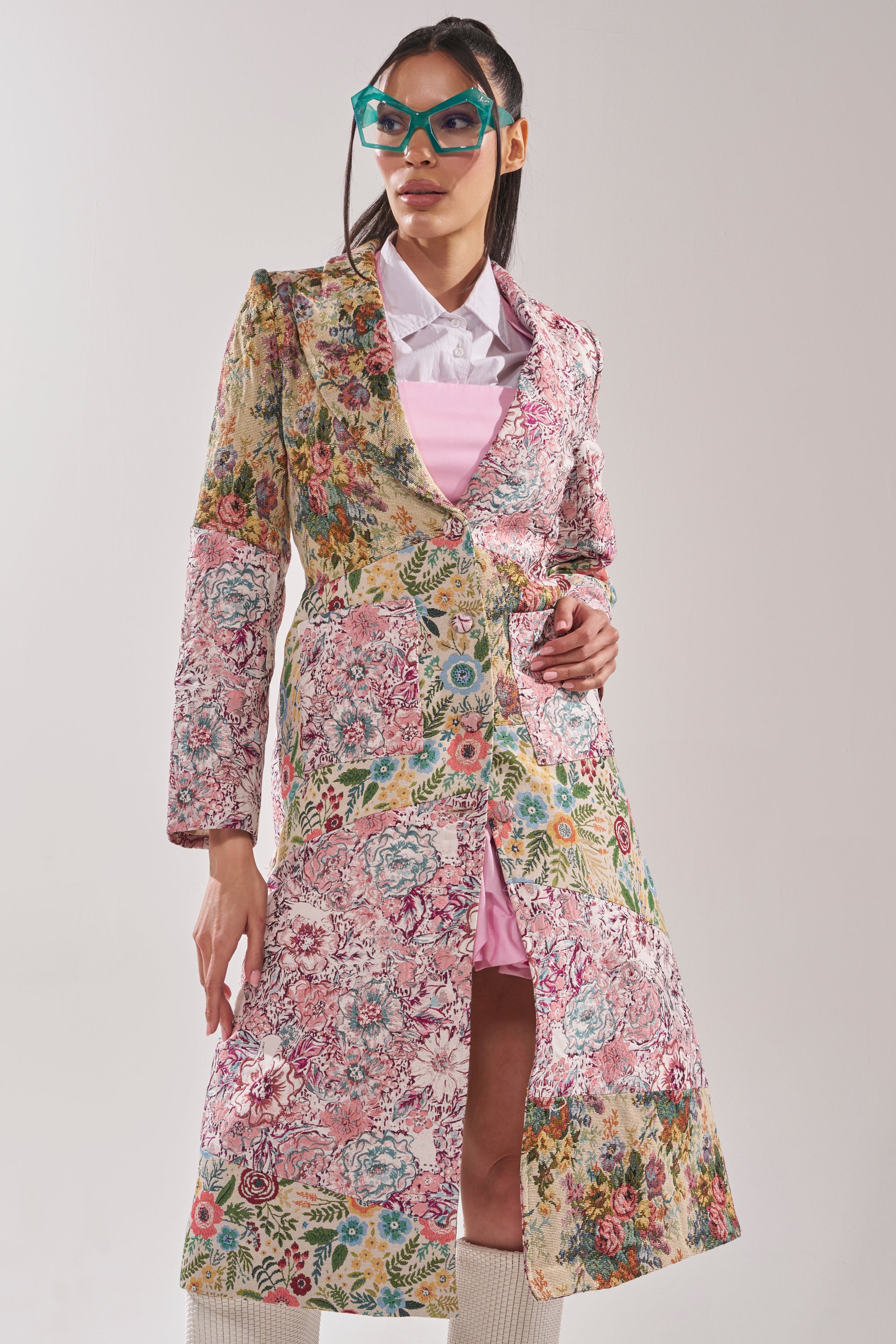 A woman in large green glasses, a white collared shirt, a pink dress, and the HAD ME AT HELLO PATCHWORK TRENCH with floral patterns stands against a plain light background. 1