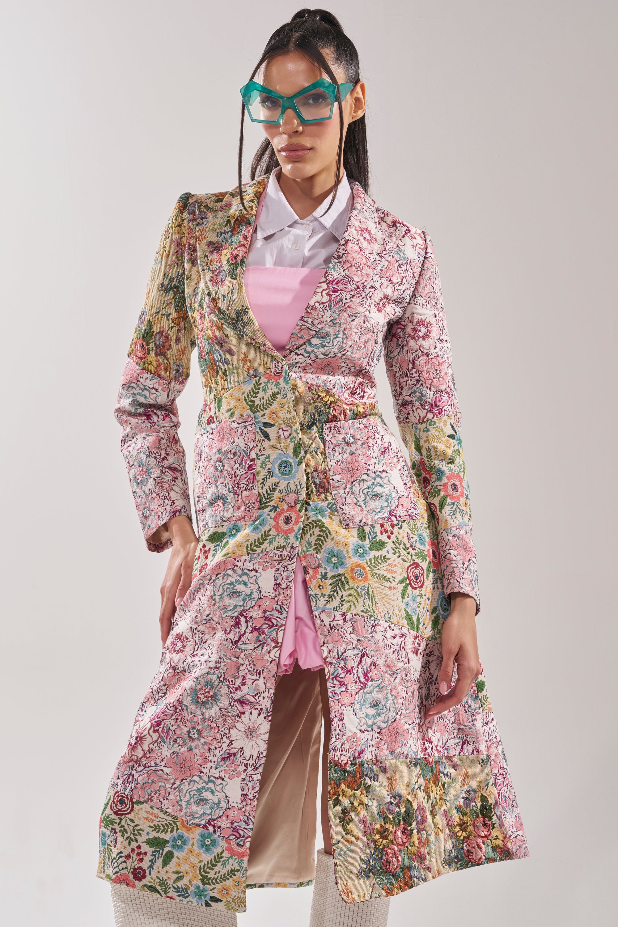Wearing the HAD ME AT HELLO PATCHWORK TRENCH over a pink dress and white shirt, paired with teal angular glasses, a person poses confidently against a plain background. 2