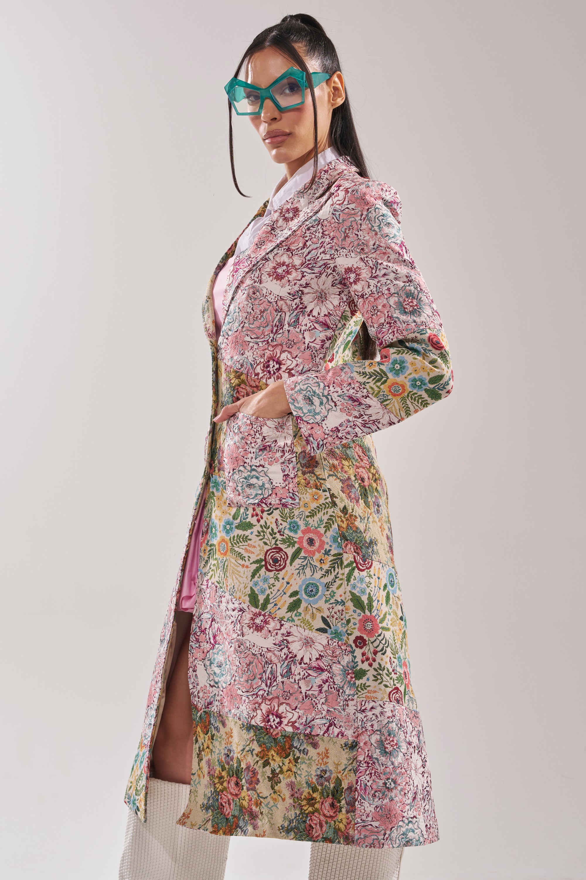 A woman poses confidently in the HAD ME AT HELLO PATCHWORK TRENCH, paired with teal glasses and knee-high white boots, hands in pockets, standing against a plain light background. 3