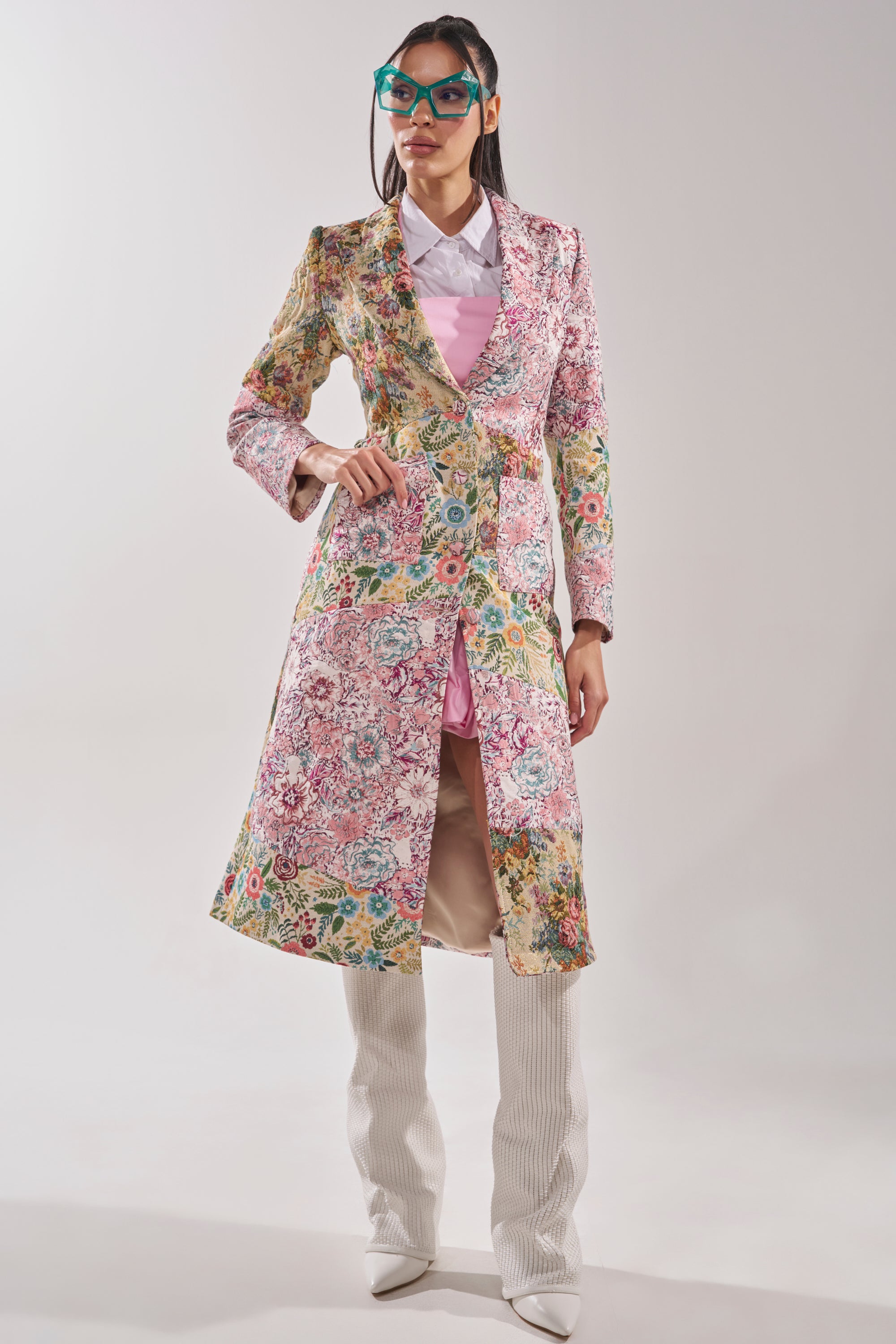 A woman models the HAD ME AT HELLO PATCHWORK TRENCH over a pink dress, white collared shirt, and tall white boots, accessorized with blue cat-eye sunglasses as she poses confidently against a plain background. 5