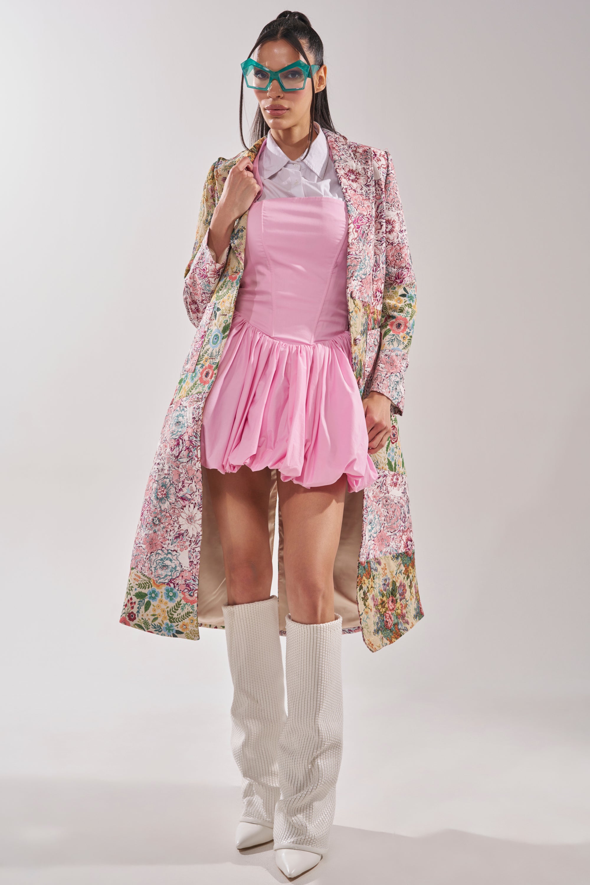 A woman stands confidently against a plain light background, wearing teal glasses, a white collared shirt, a light pink mini dress, and the HAD ME AT HELLO PATCHWORK TRENCH with white knee-high textured boots. 6