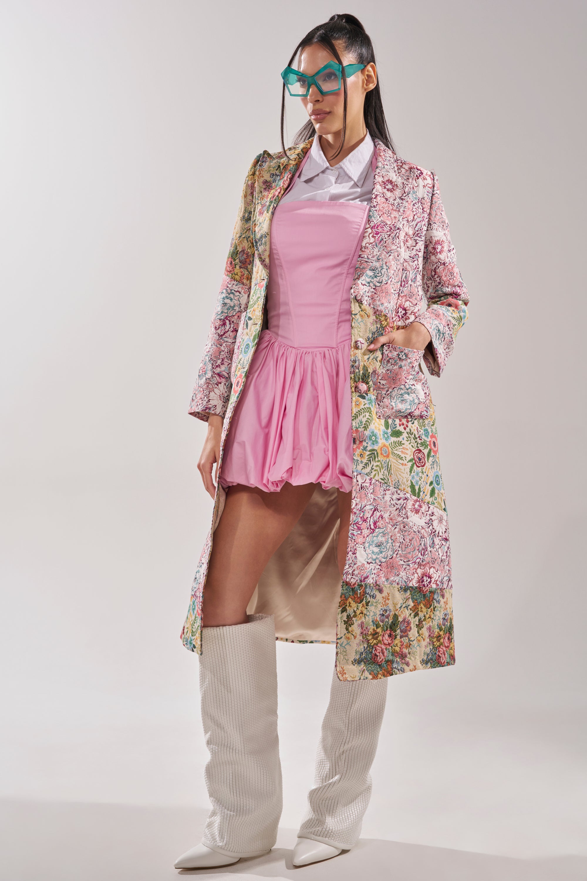 A woman poses in the HAD ME AT HELLO PATCHWORK TRENCH over a pink dress with a white collar, knee-high white boots, and blue statement glasses against a plain light background. 7
