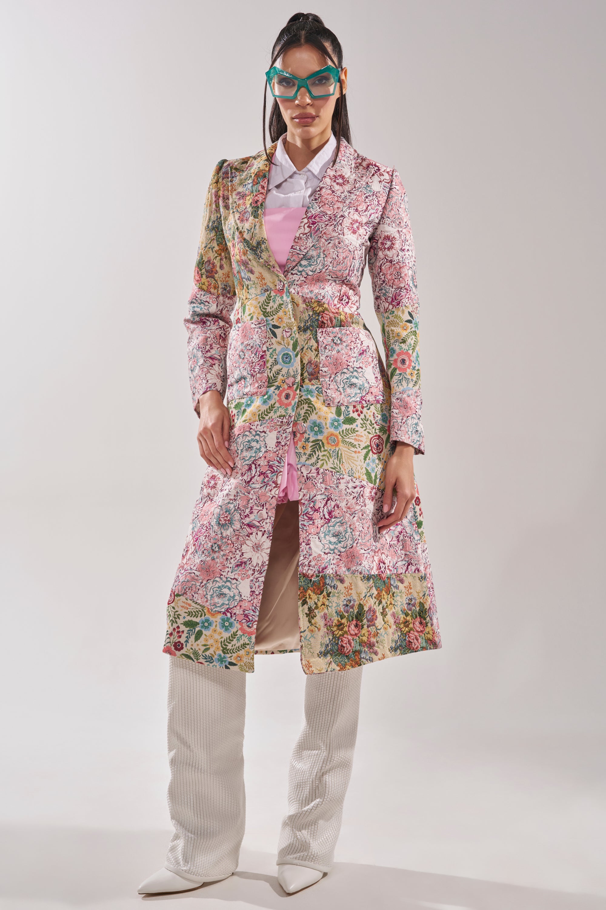A woman wears blue-green glasses and the HAD ME AT HELLO PATCHWORK TRENCH over a pink dress shirt, paired with white textured knee-high boots, as she stands facing forward against a plain light background. 8