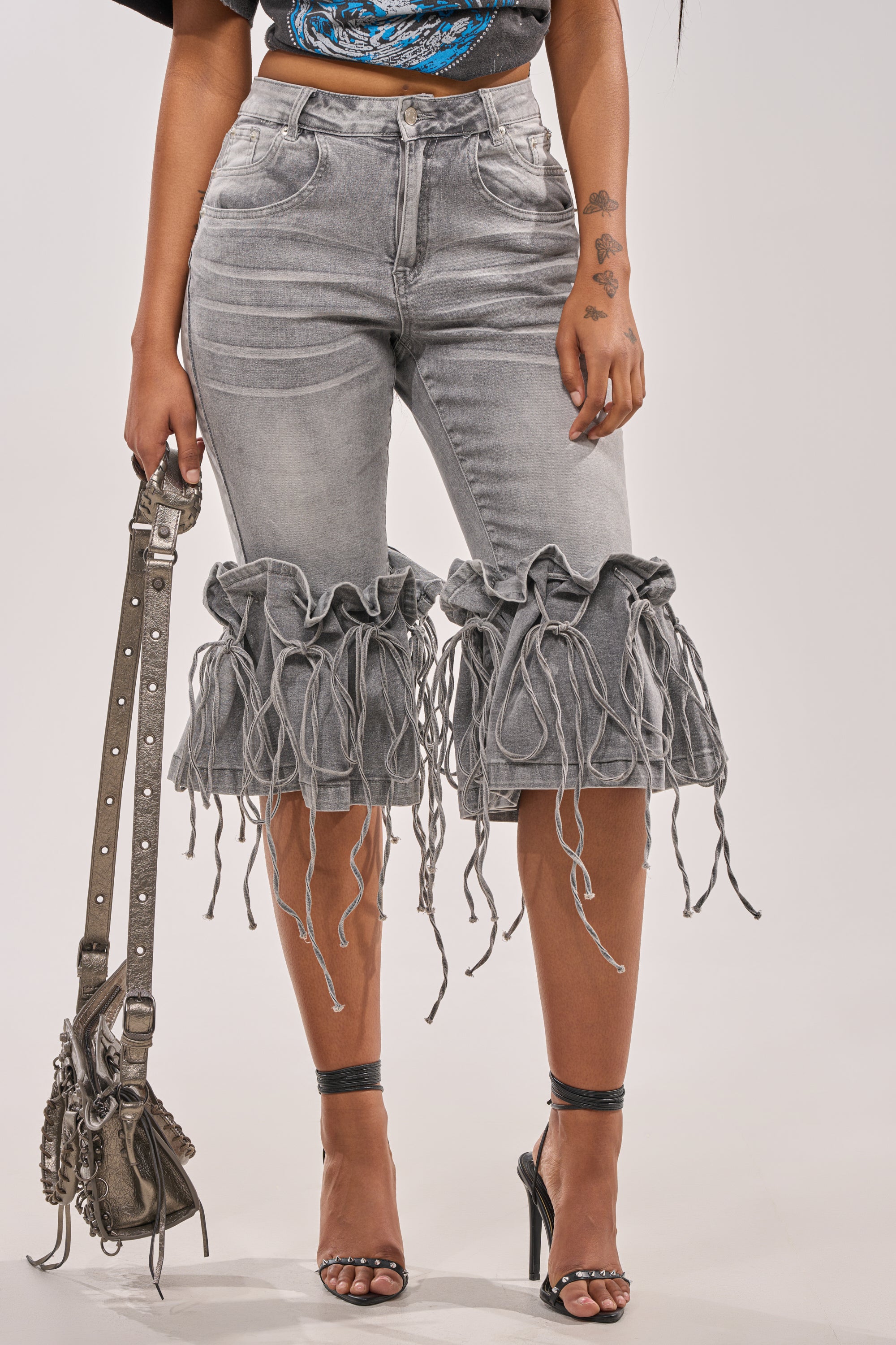 A person wears the JUMP ON IT DENIM BERMUDA SHORT in light gray with a ruffled, frayed hem, paired with black high-heeled sandals, a patterned black top, and carries a studded gray handbag. 0