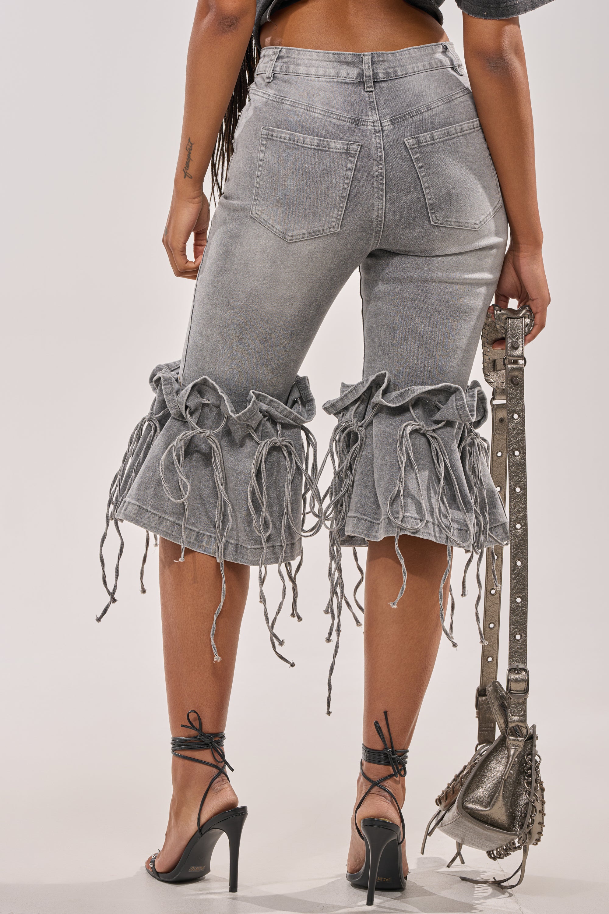 A person faces away wearing the JUMP ON IT DENIM BERMUDA SHORT with frayed ruffle knee details, paired with black strappy heels and a silver studded handbag. 5