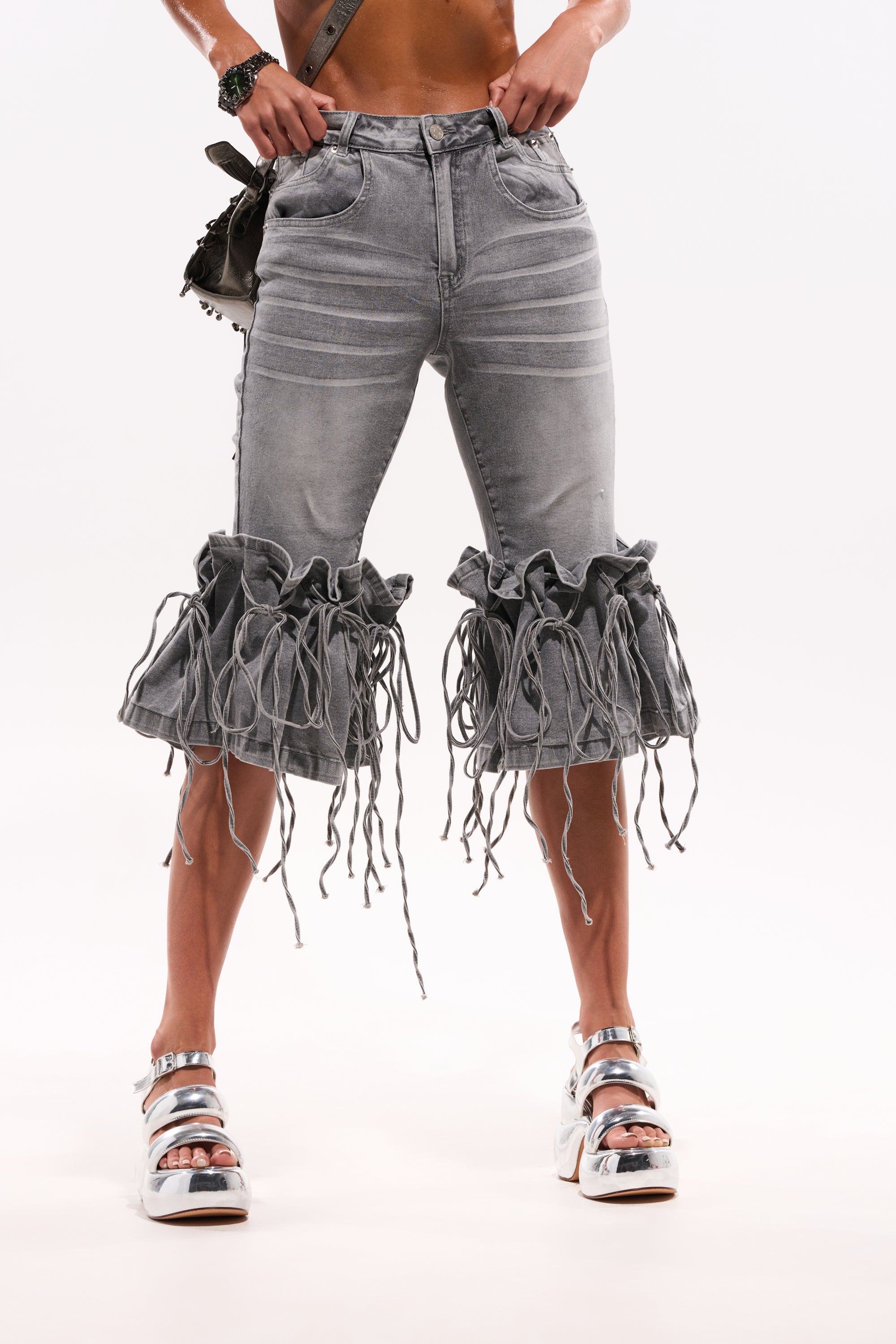 Wearing the JUMP ON IT DENIM BERMUDA SHORT in light gray with ruffle hem and fringe details, paired with silver platform sandals and a dark bag, this person stands against a plain white background. 3