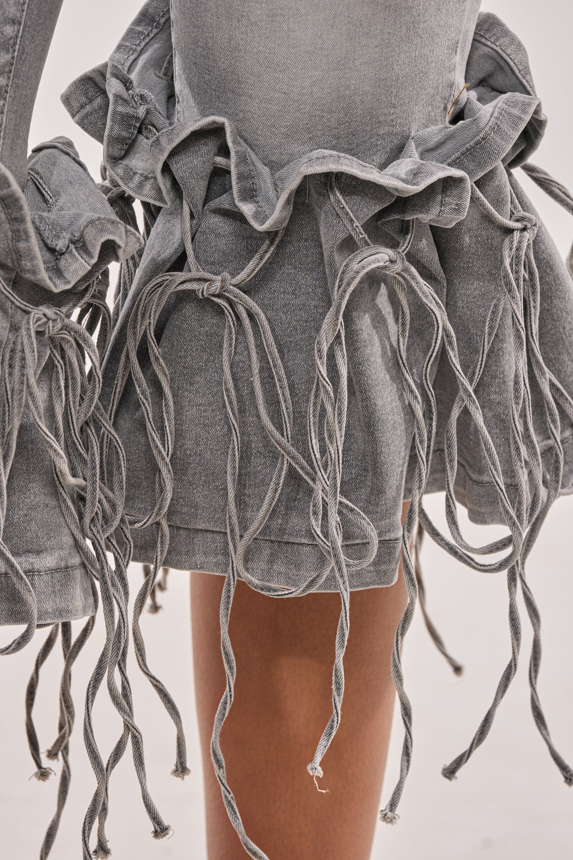 Close-up of a person wearing the JUMP ON IT DENIM BERMUDA SHORT, featuring high-rise fit, ruffled edges, and multiple loose twisted fabric cords for a textured layered look. The neutral background is softly blurred. 7
