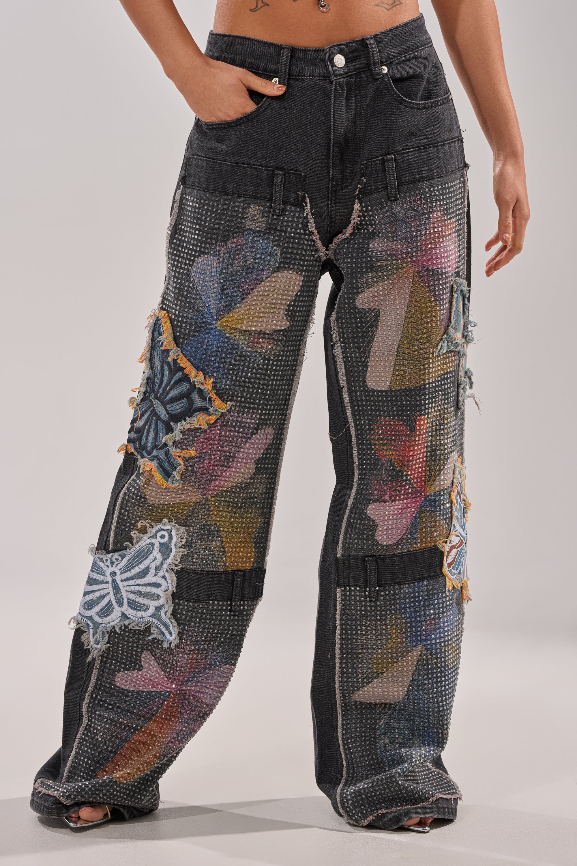A person wears BUTTERFLY EFFECT RELAXED DENIM: loose-fitting, high-waisted jeans with colorful mesh patches, large butterfly appliqués, frayed seams, and sparkling rhinestone embellishments on black denim. 0