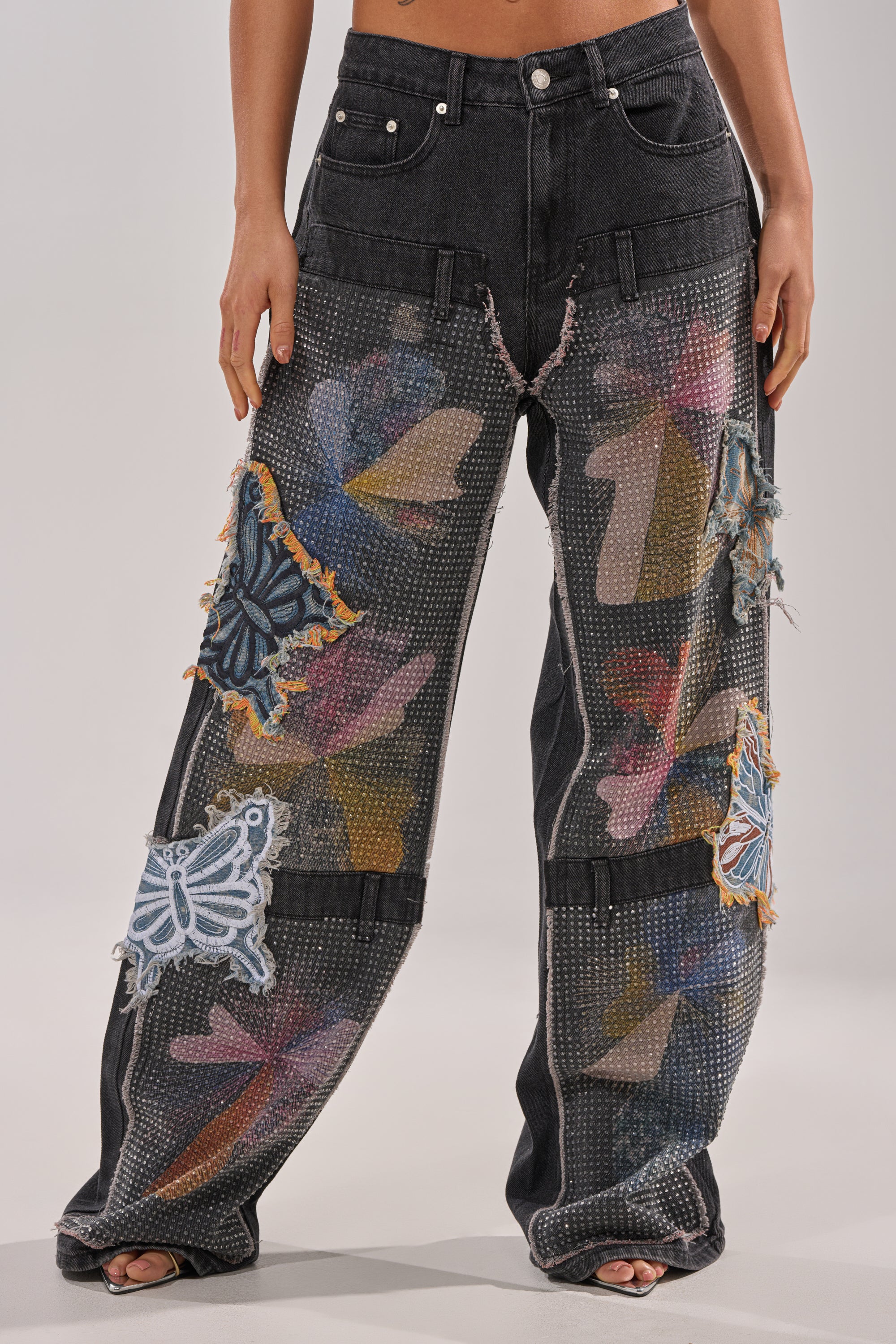 Someone wears the BUTTERFLY EFFECT RELAXED DENIM, featuring bedazzled black wide-leg jeans with sequin mesh, butterfly patches, multicolored abstract patterns, a high waist, distressed details, and goes barefoot. 1