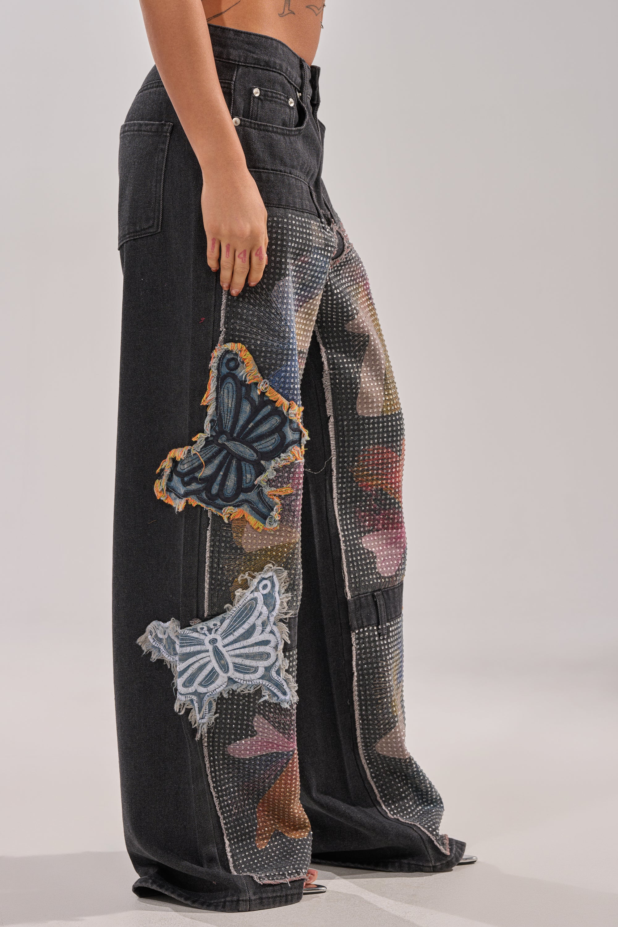 A person wears the BUTTERFLY EFFECT RELAXED DENIM, featuring large butterfly patches and mesh details, standing barefoot against a neutral backdrop with only their lower body shown. 2