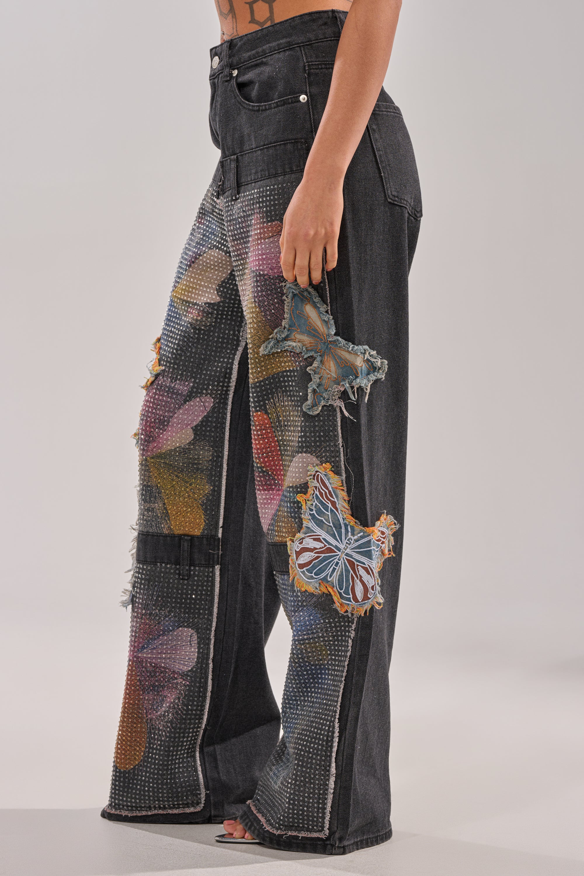 A person wearing the BUTTERFLY EFFECT RELAXED DENIM, featuring bedazzled black denim with colorful butterfly patches and rhinestones, stands sideways against a plain light background. 3