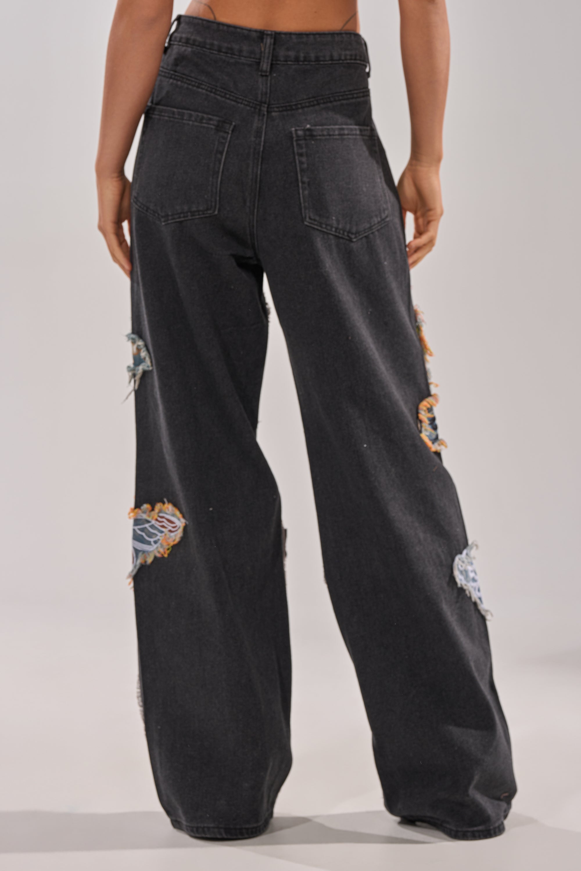 A person wearing BUTTERFLY EFFECT RELAXED DENIM—high-waisted, loose-fitting black jeans with bedazzled details, distressed patches, and frayed edges—shown from the back against a neutral background. 4