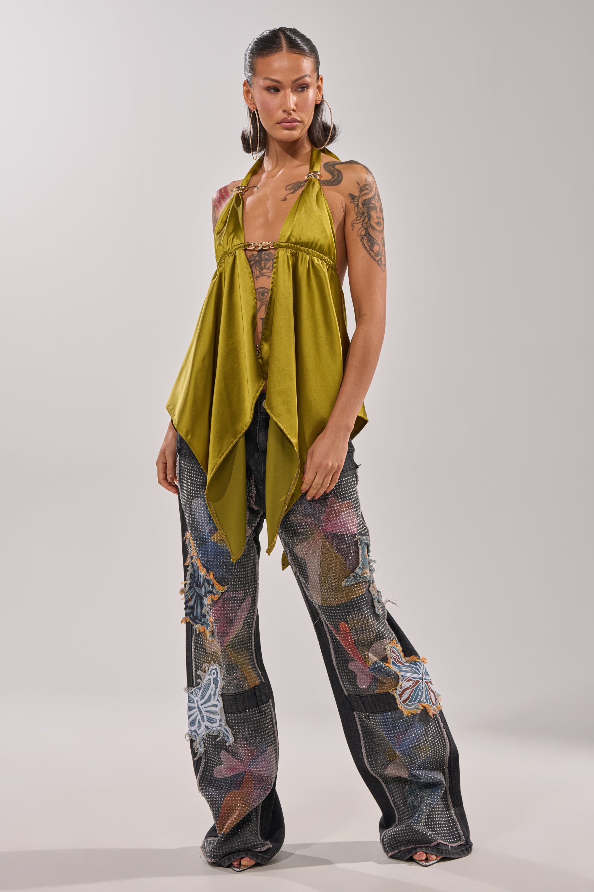 A woman models the BUTTERFLY EFFECT RELAXED DENIM with colorful patchwork and appliqués, paired with a halter-neck olive green top, open-toe shoes, hoop earrings, and shows her long hair and visible tattoos. 5