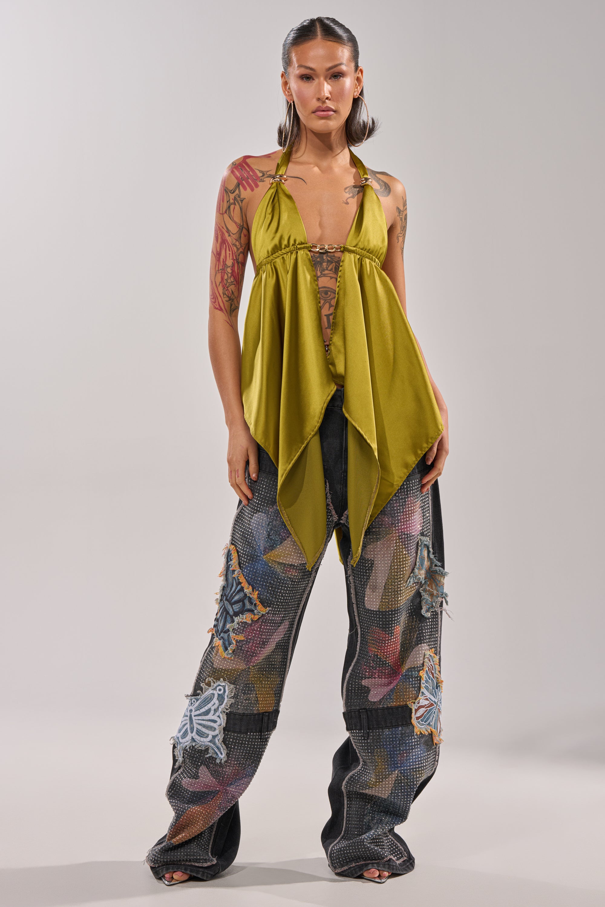 A woman with tattoos and styled brown hair poses against a light background, wearing an olive-green halter top and the BUTTERFLY EFFECT RELAXED DENIM featuring colorful patchwork and abstract butterfly-inspired designs. 6