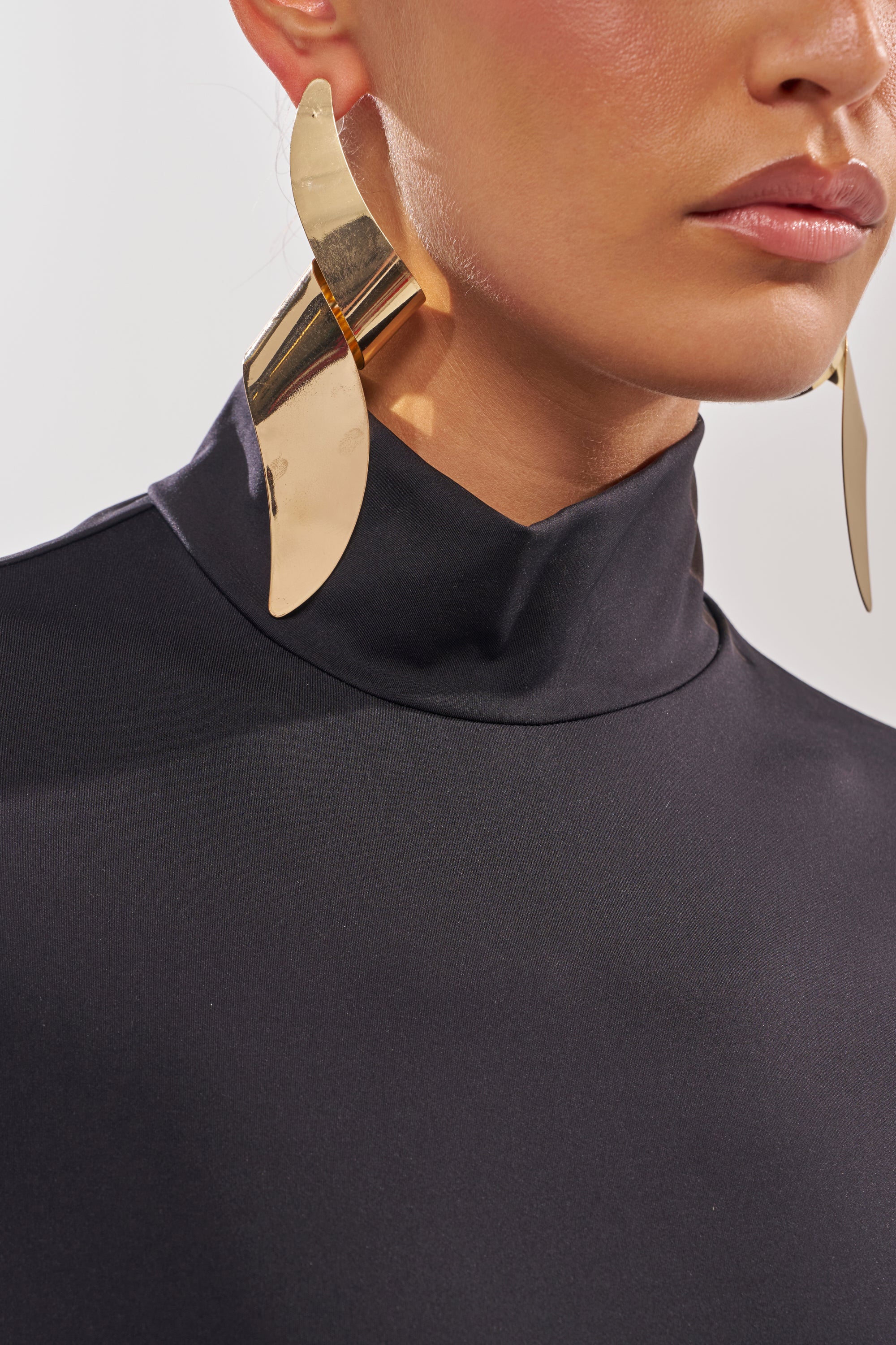 A close-up shows a woman wearing the BLACK OUT MOCK NECK OPEN BACK BODYSUIT and large, abstract gold leaf-shaped earrings, with only the lower half of her face and upper body visible. 9