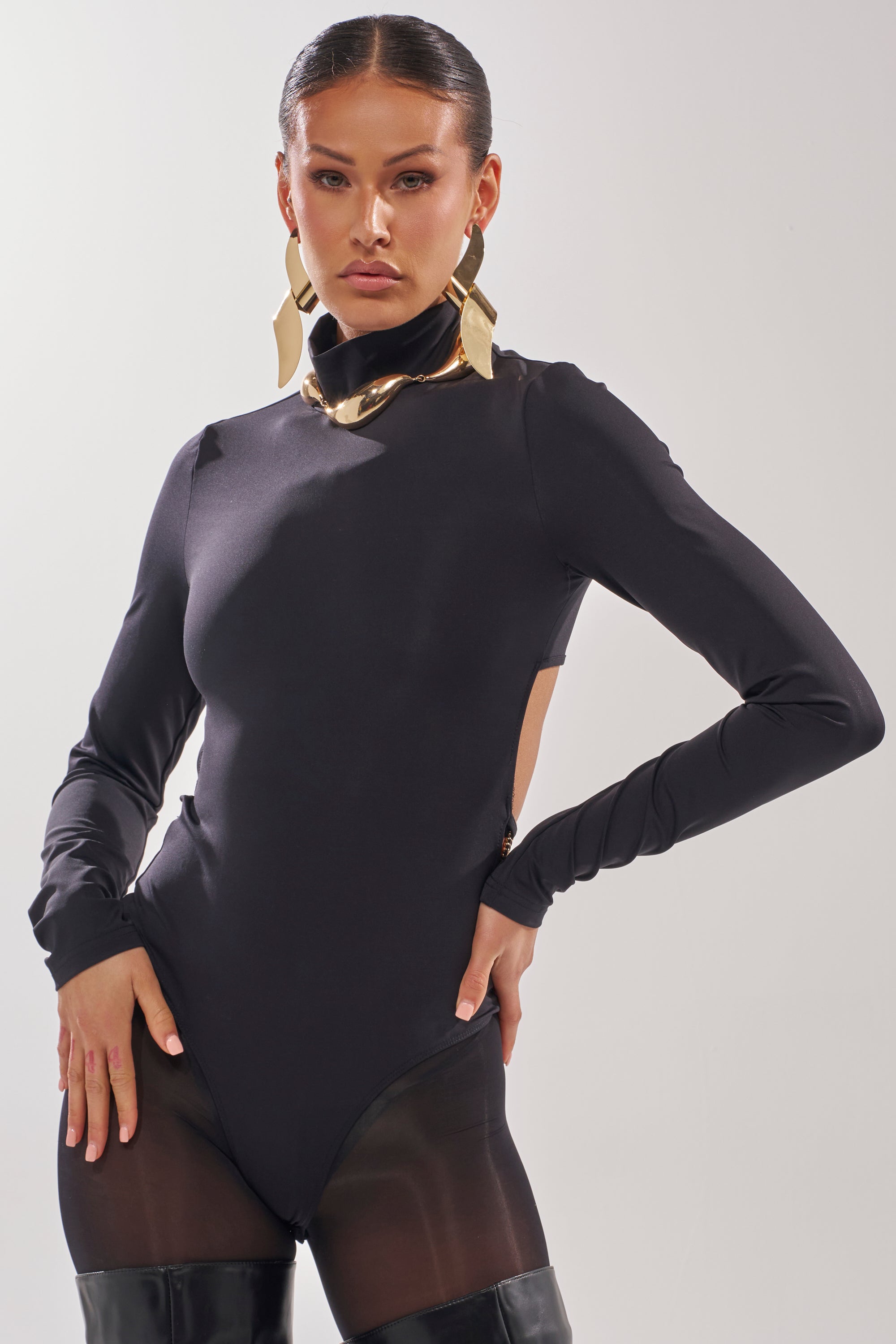 A woman strikes a pose in the BLACK OUT MOCK NECK OPEN BACK BODYSUIT, styled with bold gold earrings and black thigh-high boots against a simple backdrop. 2