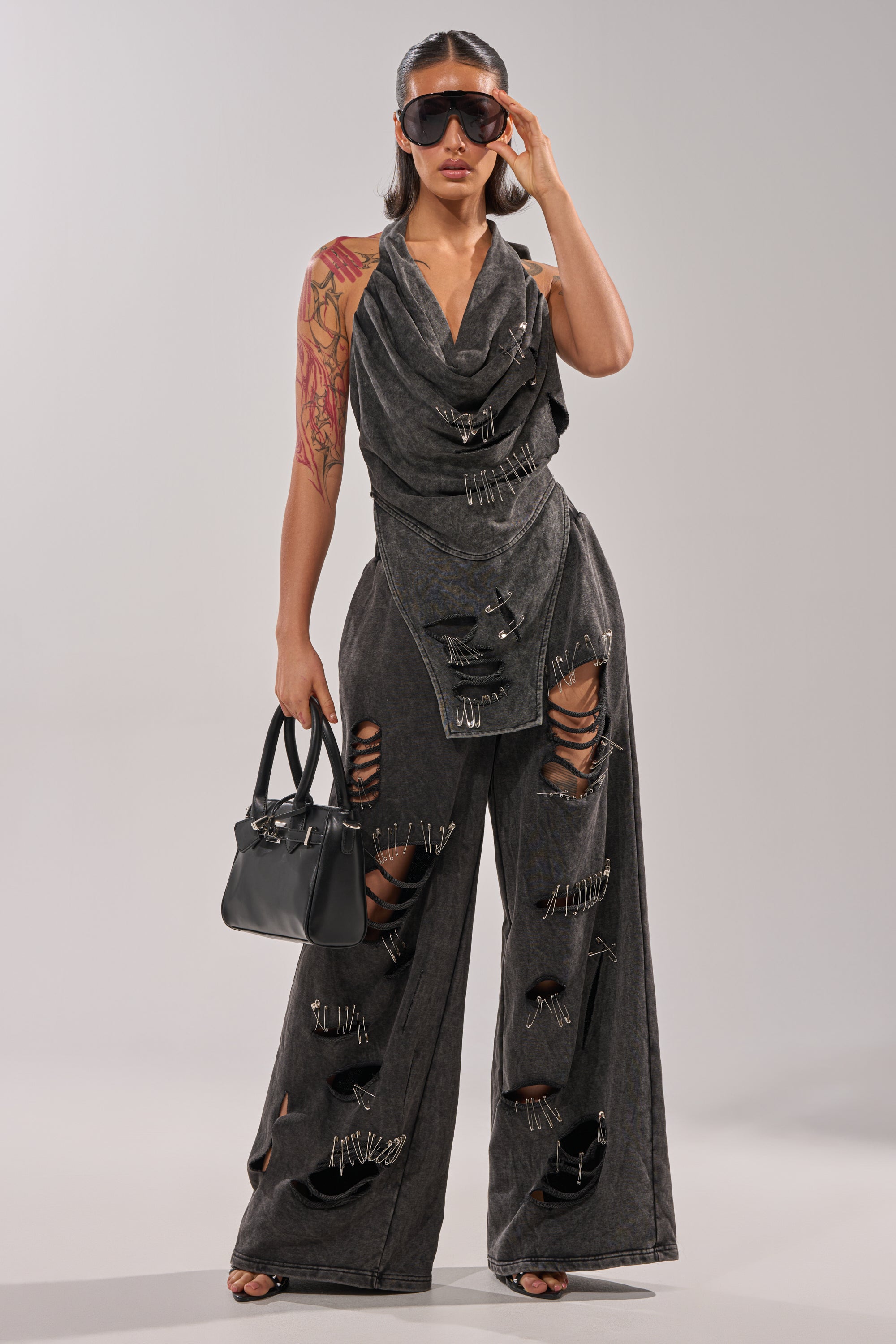 A woman in large sunglasses and visible arm tattoos stands against a plain light background, wearing grunge streetwear: the LEILA DISTRESSED WIDE LEG JOGGER in dark gray with safety-pin accents, paired with a black handbag. 0