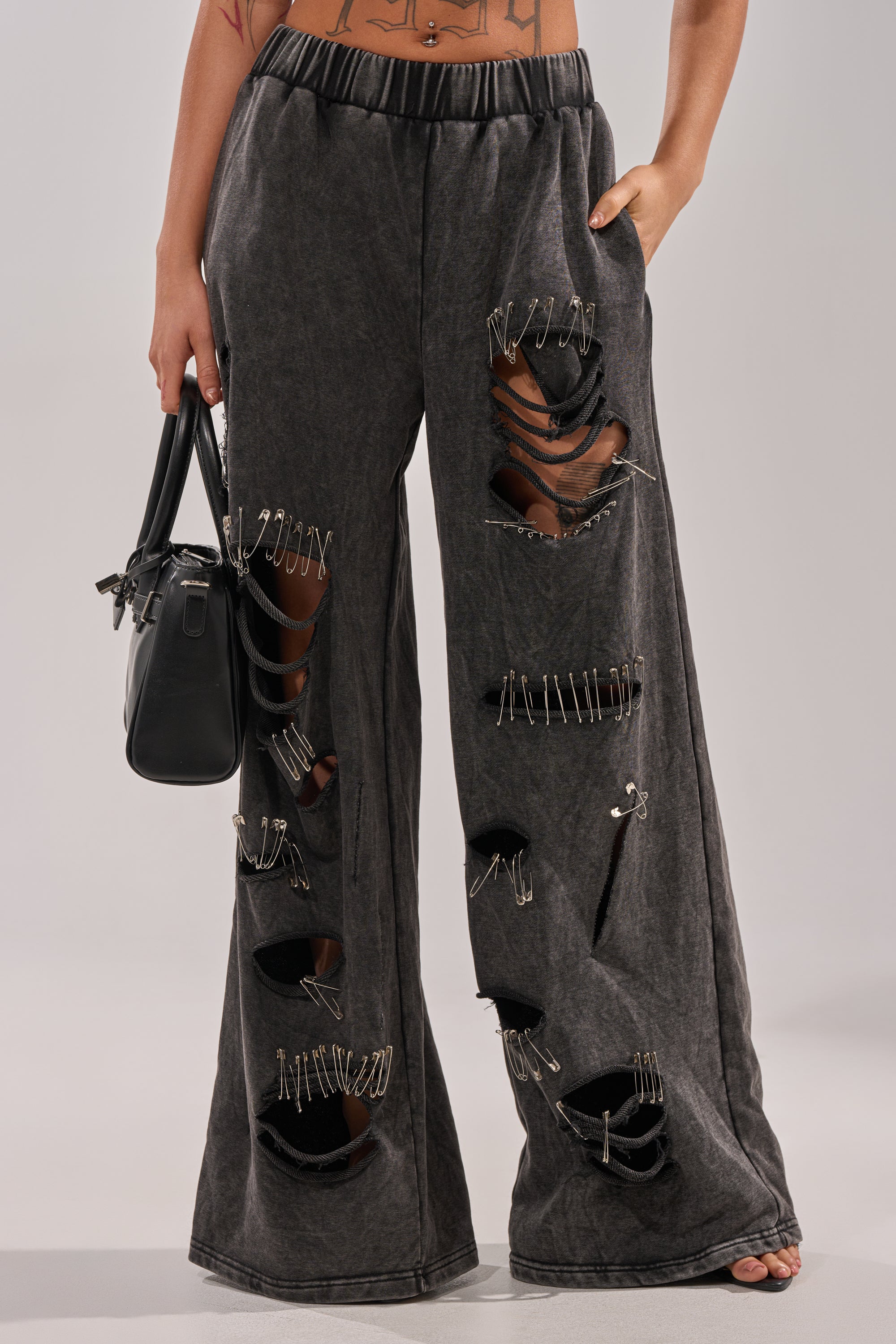 A person wears LEILA DISTRESSED WIDE LEG JOGGER: loose, black joggers with bold rips and safety-pin accents, paired with a black handbag. Only their lower torso and legs are visible in the image. 2