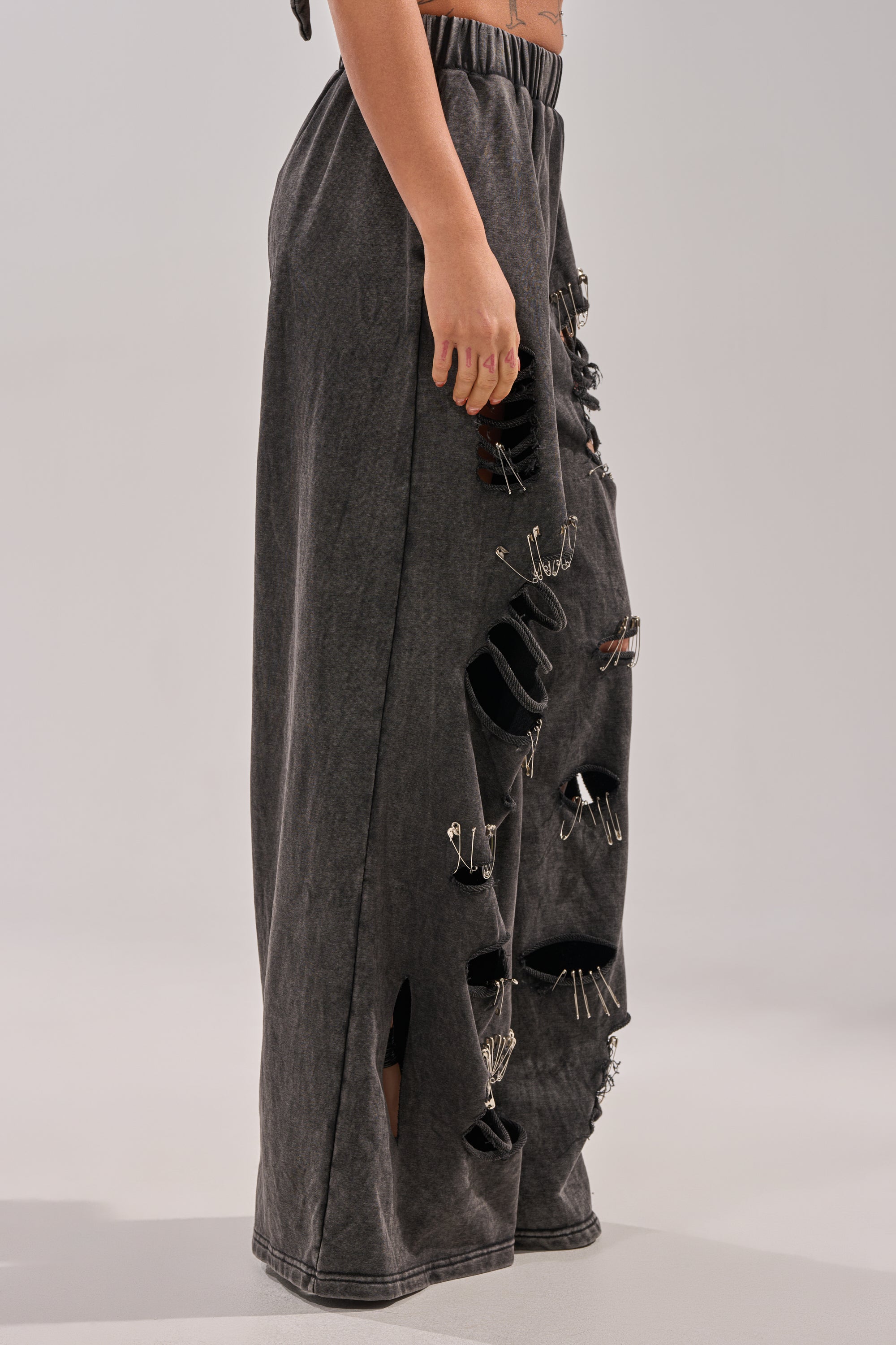 Model displays the LEILA DISTRESSED WIDE LEG JOGGER, featuring dramatic rips and safety-pin details for a grunge streetwear vibe. Only the lower half of the body is shown, set against a plain background. 3