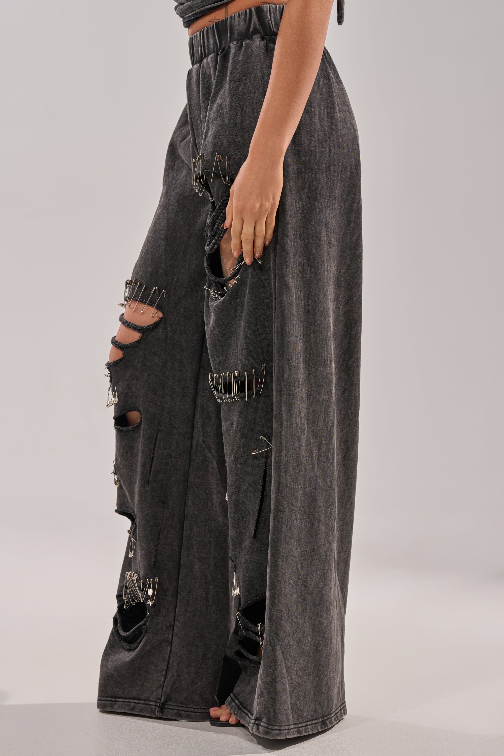 A person showcases the LEILA DISTRESSED WIDE LEG JOGGER—black, wide-leg jeans with major rips and safety-pin details—standing barefoot against a plain light backdrop. 4
