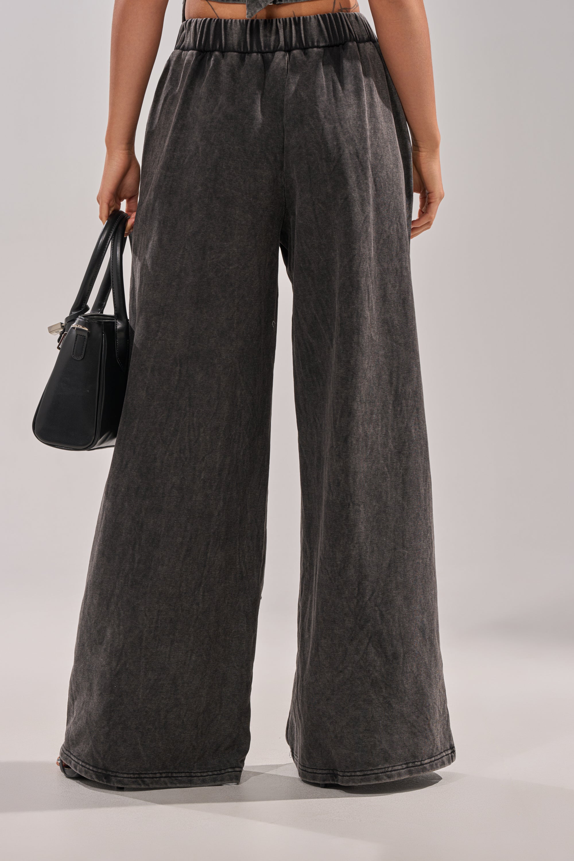A person faces away, wearing the LEILA DISTRESSED WIDE LEG JOGGER and holding a black handbag in their right hand. Their top is not visible, and the background is neutral and plain. 5