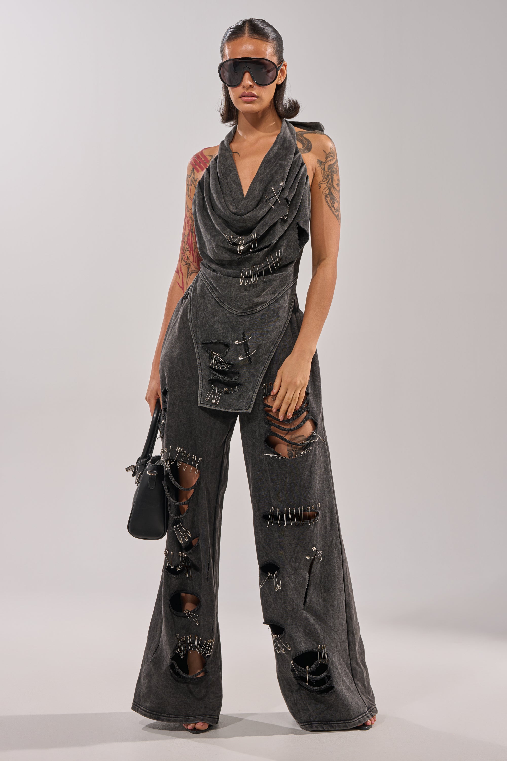 A woman in grunge streetwear wears oversized sunglasses and the LEILA DISTRESSED WIDE LEG JOGGER, featuring large rips and safety-pin accents. She carries a black handbag, showing off her colorful tattoo against a plain background. 7