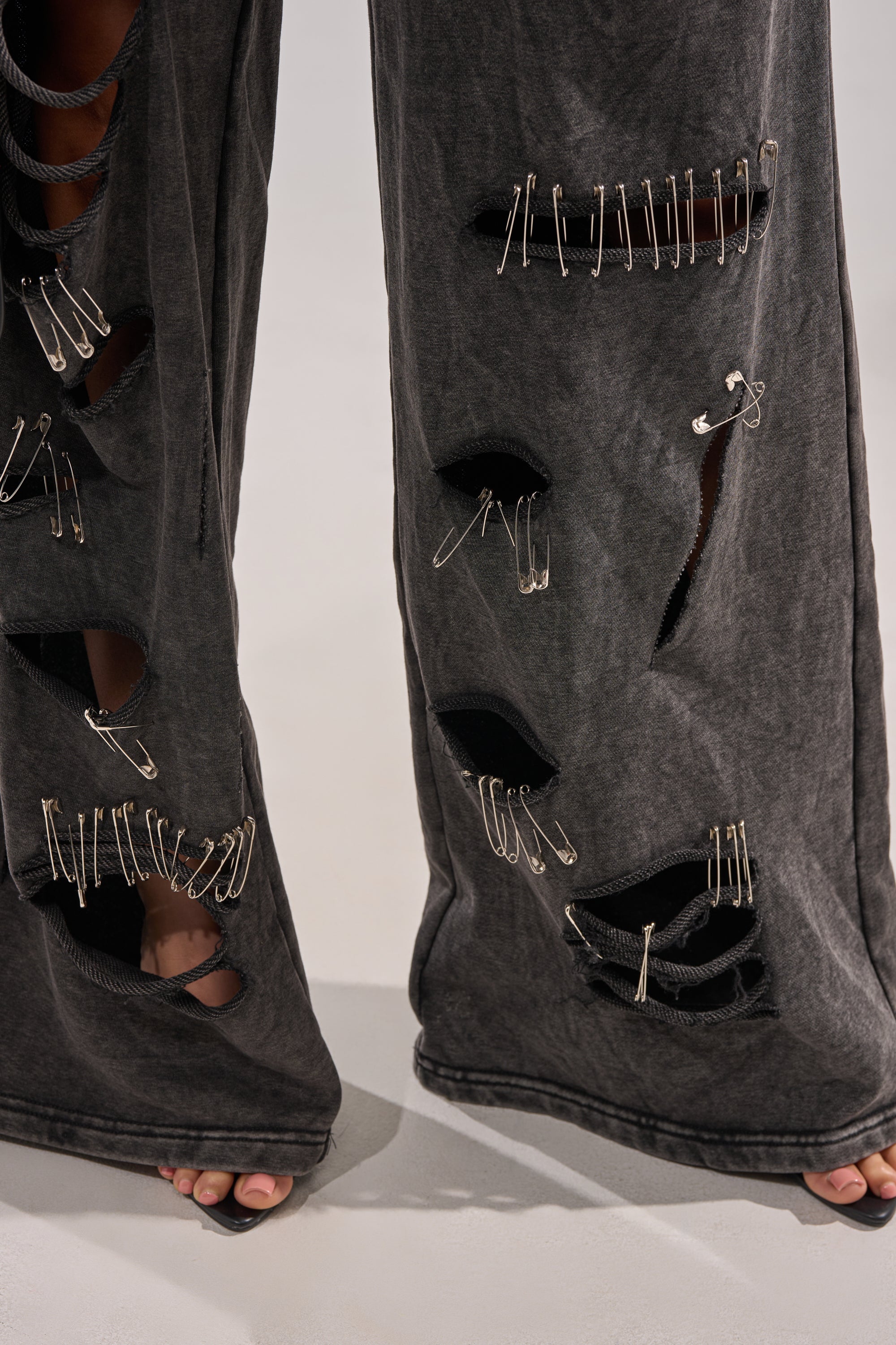 Close-up of the LEILA DISTRESSED WIDE LEG JOGGER, featuring large, irregular holes and safety-pin details. The model stands barefoot on a light surface, toes peeking from the frayed hem—an edgy twist on grunge streetwear. 8