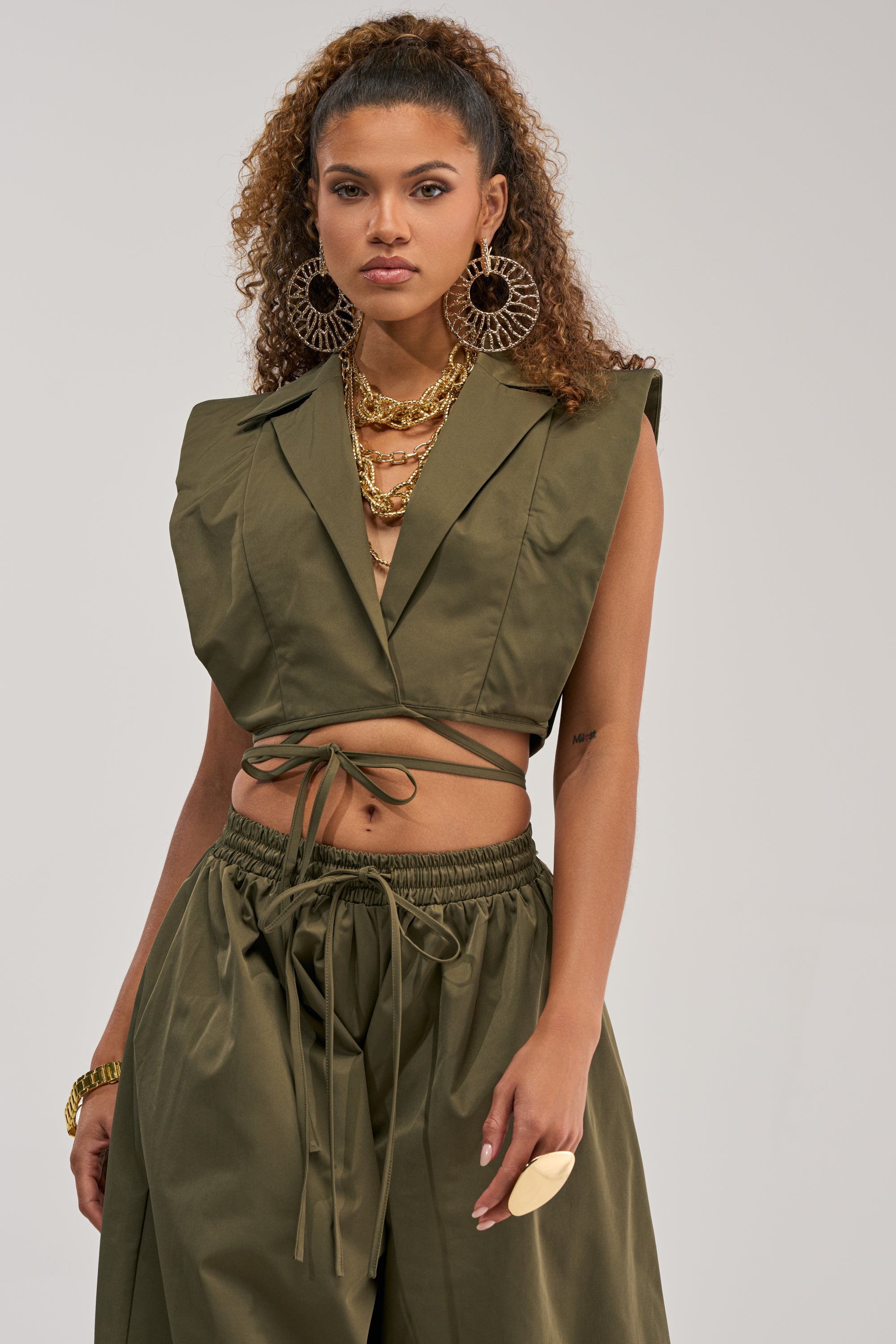A woman with curly hair wears the GUESS WHO VNECK COLLARED BLOUSE IN OLIVE and matching wide-leg pants, styled with hoop earrings, layered gold necklaces, a chunky bracelet, and a statement ring.