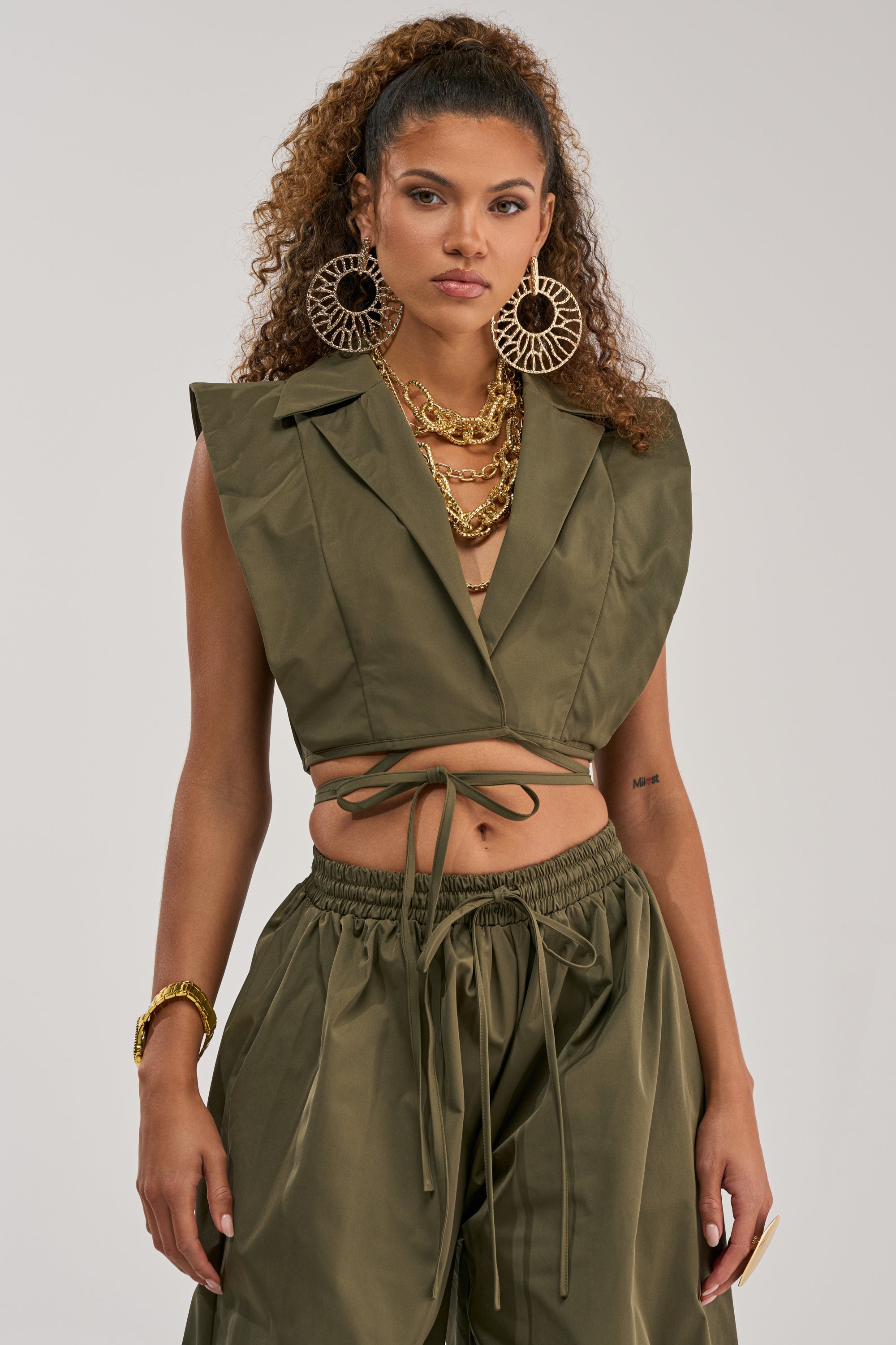 A woman wears the GUESS WHO VNECK COLLARED BLOUSE IN OLIVE paired with matching wide-leg pants, embodying modern power dressing. She accessorizes with large statement earrings, layered gold necklaces, and a bracelet, while her curly hair is pulled back. She stands against a plain background.