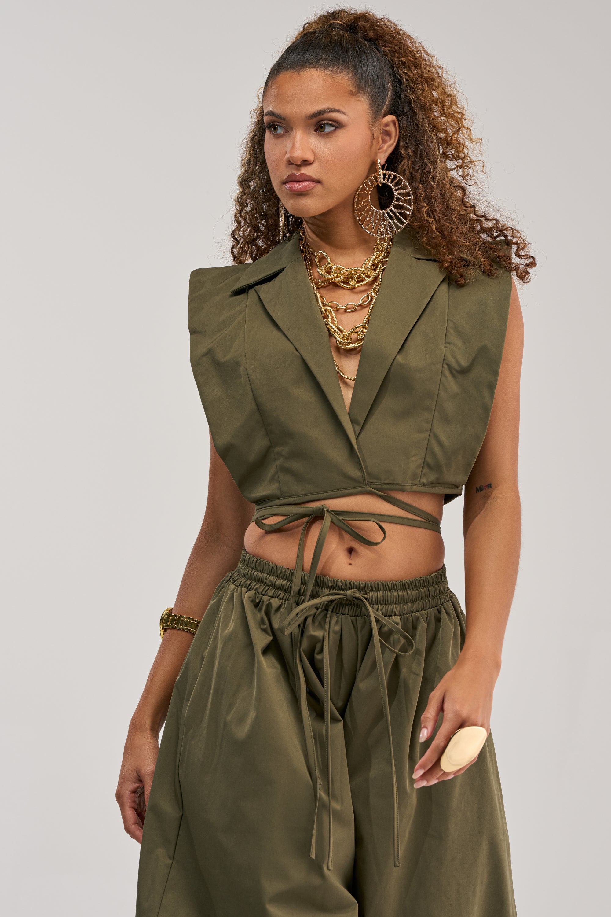 Dressed in the GUESS WHO VNECK COLLARED BLOUSE IN OLIVE and coordinating wide-leg pants, a woman channels modern power dressing with large earrings, layered necklaces, a chunky bracelet, and a statement ring as she stands against a plain light background.