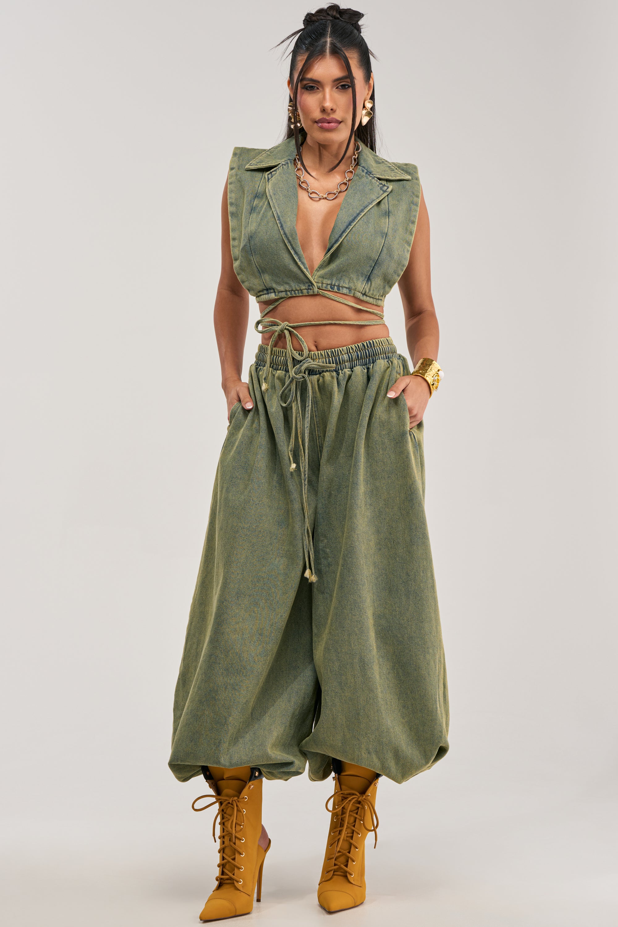 A woman stands facing forward with her hands in her pockets, wearing a sleeveless green collared cropped top, the GUESS WHO BALLOON PANT IN DENIM featuring a high-rise relaxed fit and drawstring waist, gold jewelry, and tan lace-up boots.