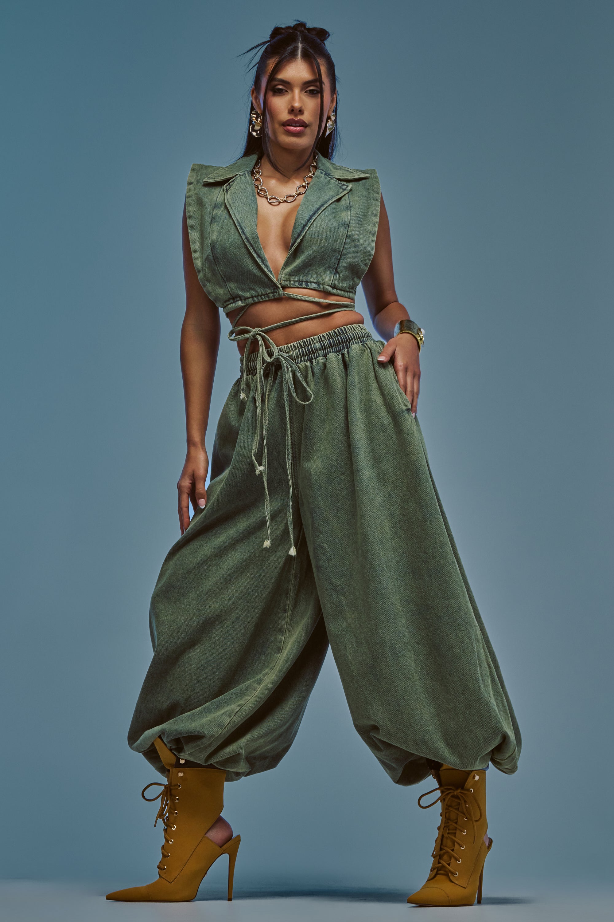 A woman models the GUESS WHO BALLOON PANT IN DENIM, styled with a sleeveless deep V-neck green crop top, mustard yellow high-heeled boots, gold jewelry, and set against a blue-gray background.